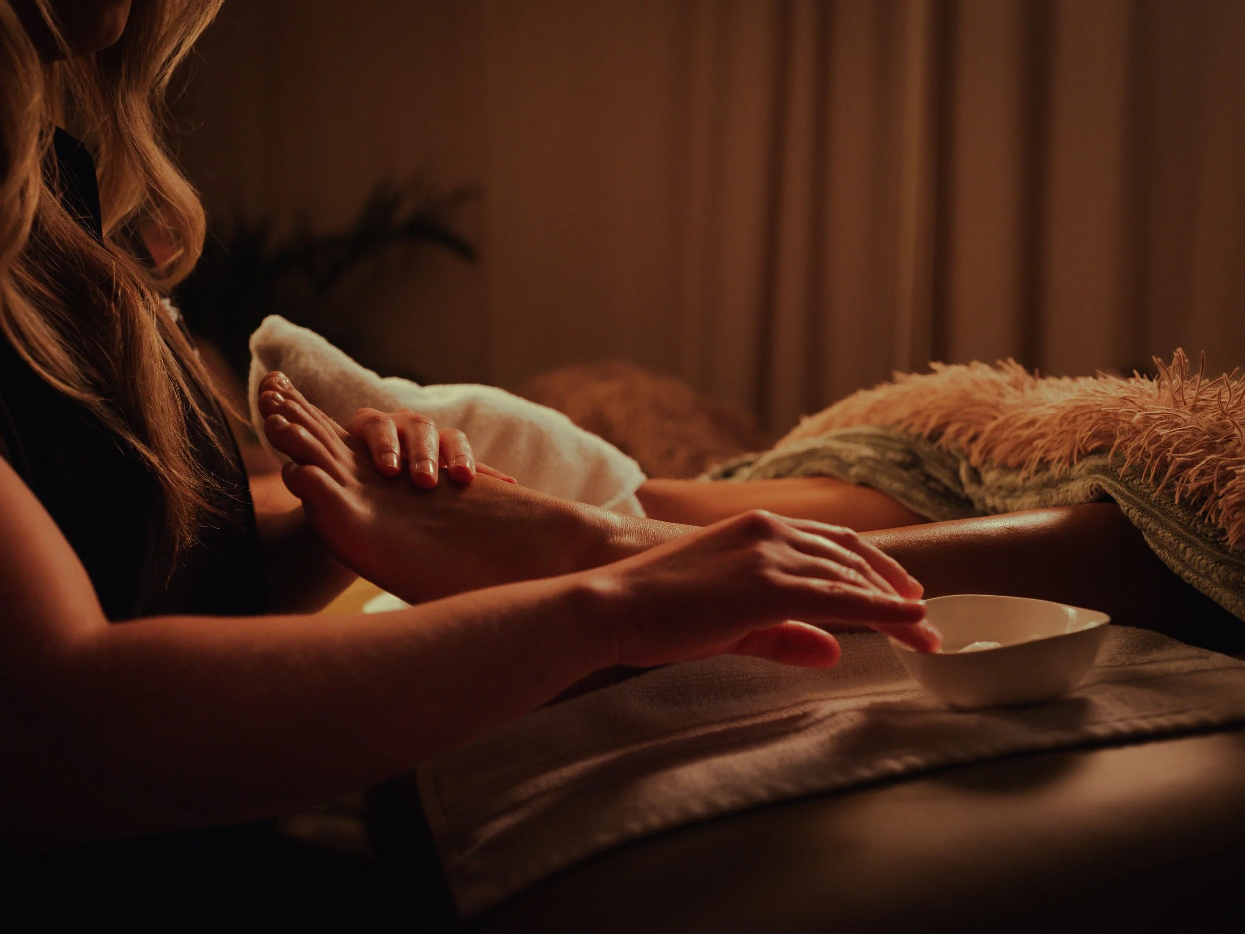 A person with long hair touching or holding a white object, possibly a book or a box, near a table with a furry or textured item on it. The background is dark with curtains.
