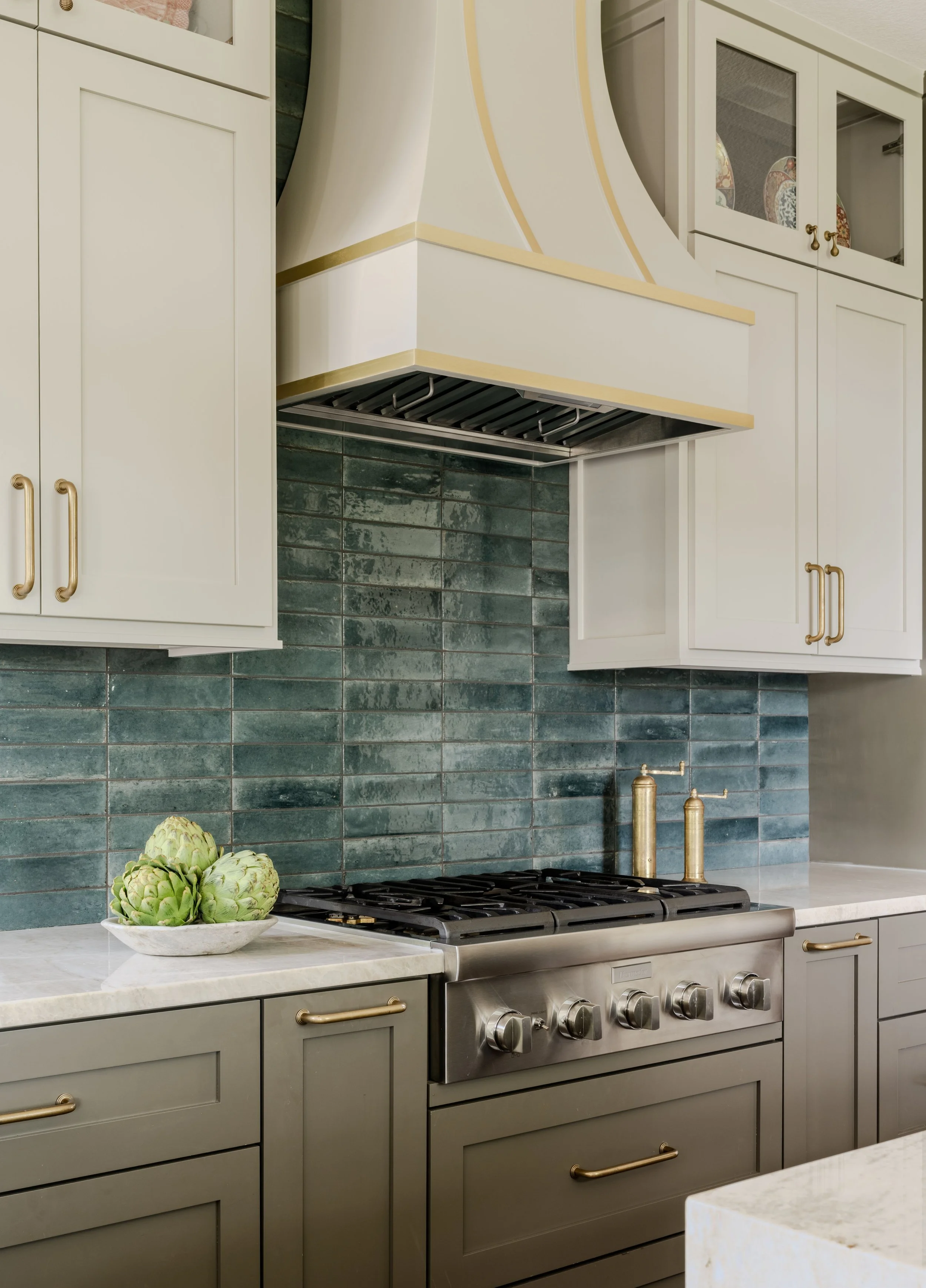 This custom kitchen at the Schug Cove project layers handcrafted blue tile, warm gray cabinetry, quartzite countertops, and a sculptural plaster range hood with brass detailing to create a refined yet welcoming gathering space. Cyndi Hopkins Design b