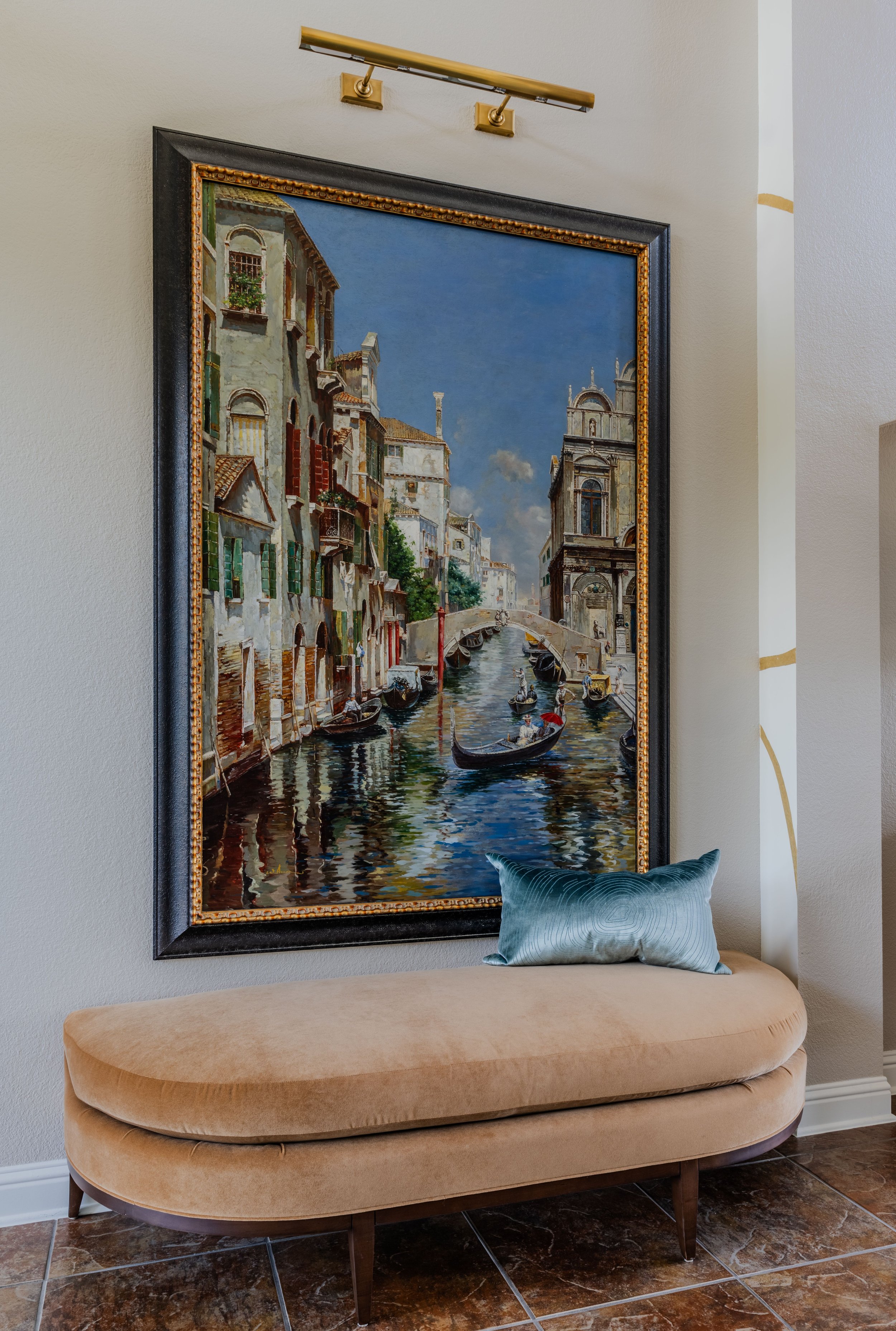 This elegant foyer vignette at the Schug Cove project pairs oversized Venetian artwork with sculptural seating and tailored lighting to create a refined entry experience. Designed by Cyndi Hopkins Design, the curved upholstered bench, brass picture l