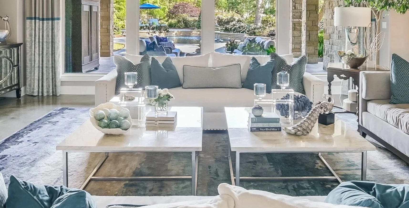 Elegant living room with white sofas, glass coffee tables, decorative pillows, and view of lush outdoor patio with pool and seating area.