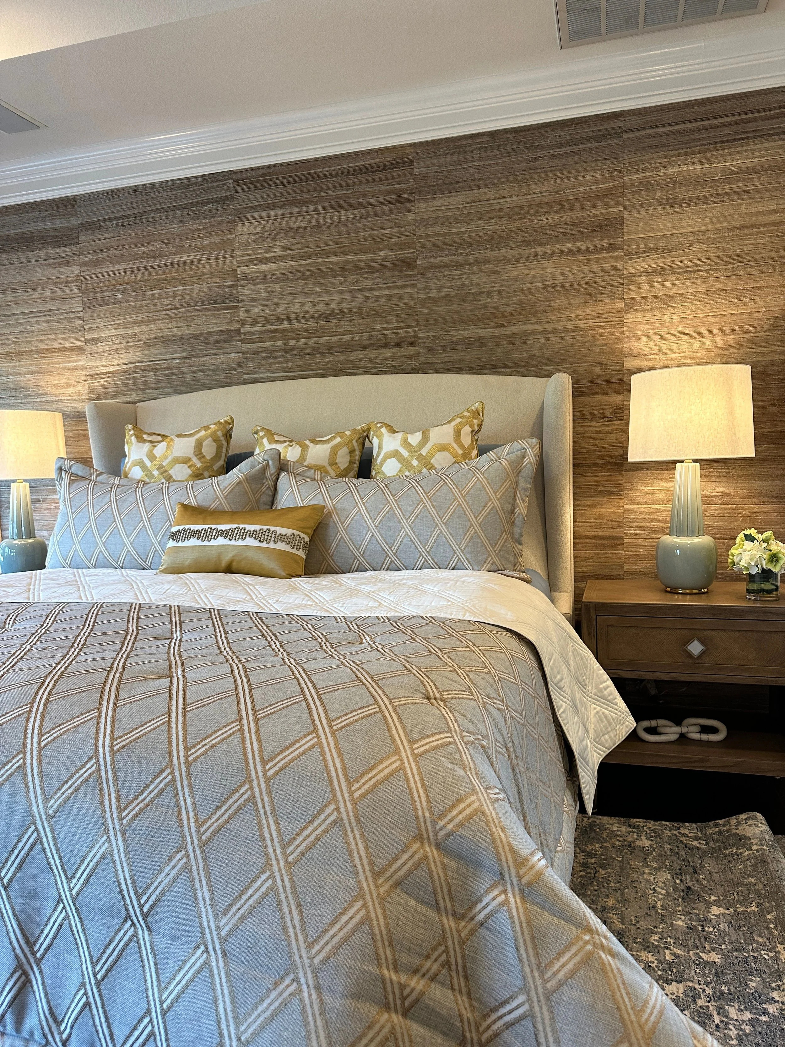 This sophisticated primary bedroom by Cyndi Hopkins Design layers warm neutrals, tailored bedding, and a textured grasscloth feature wall to create a calm and elevated retreat. Soft blue accents, rich wood furnishings, and custom textiles bring depth