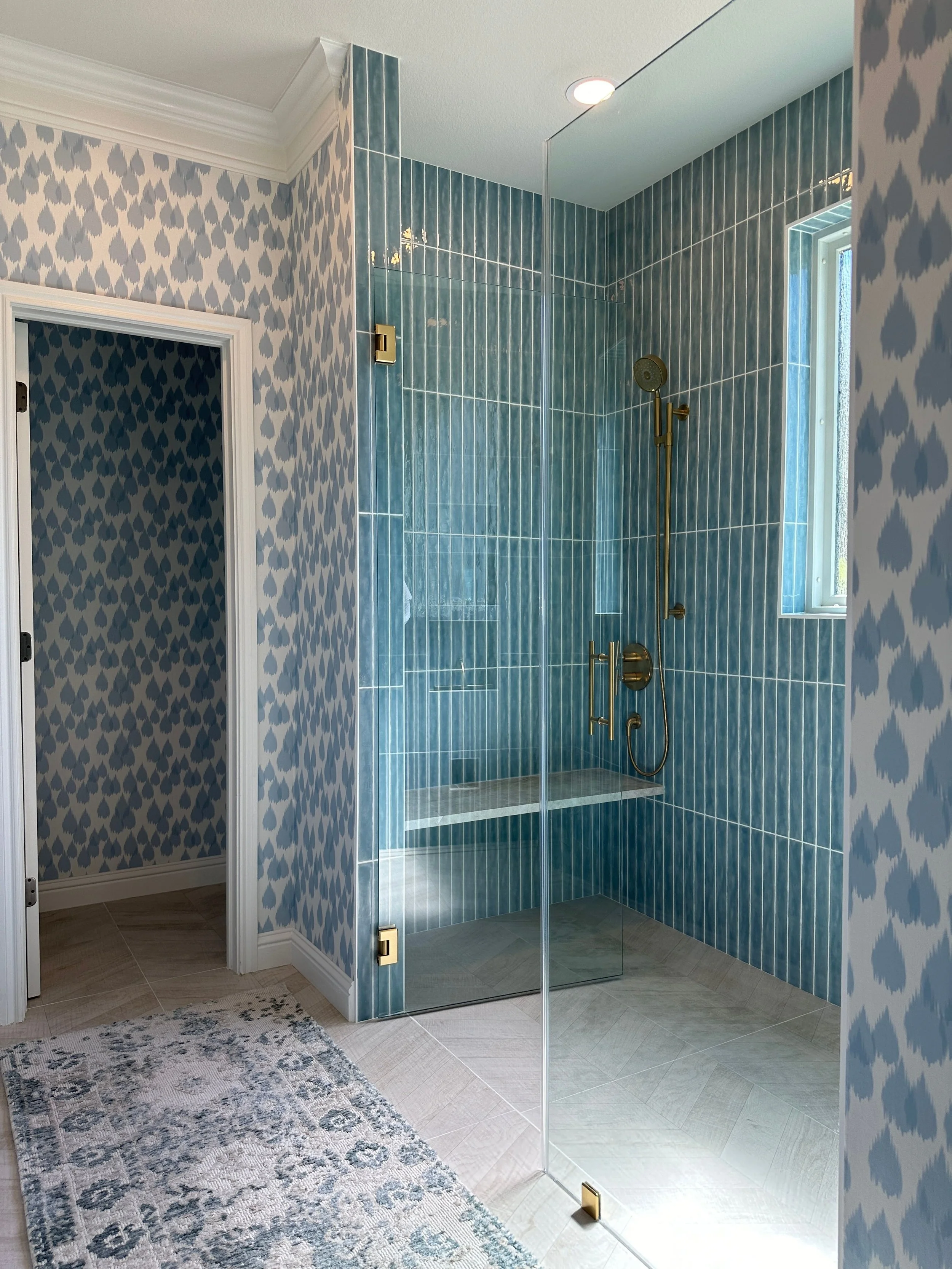 This custom bathroom at the Schug Cove project blends bold color with timeless detailing through a layered mix of patterned wallpaper, vertically stacked blue tile, warm brass plumbing fixtures, and a frameless glass shower enclosure. Designed by Cyn