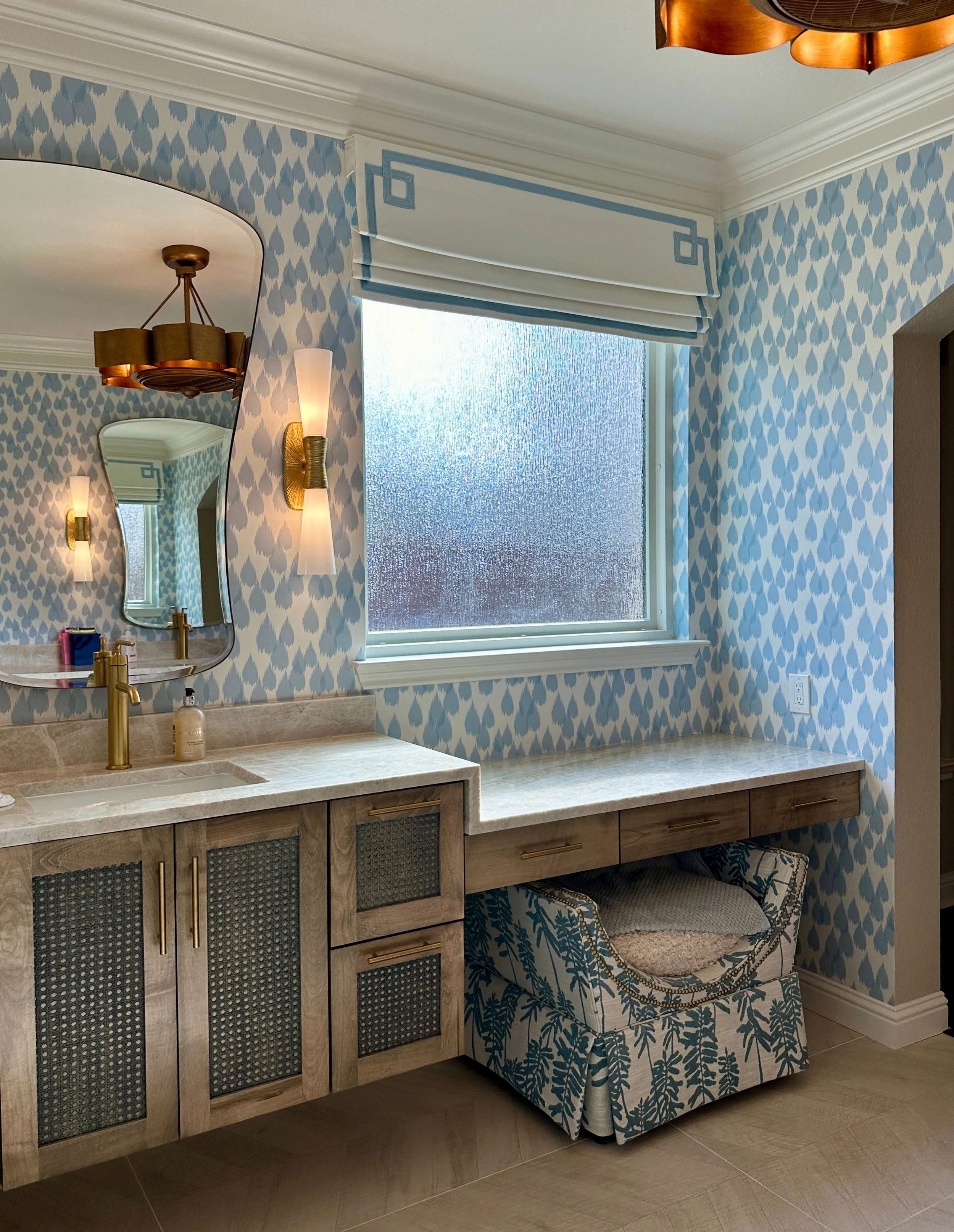This custom bathroom by Cyndi Hopkins Design combines soft blue patterned wallpaper, warm wood cabinetry, woven texture details, and brass lighting for a refined and inviting retreat. The built in makeup vanity and layered finishes create a highly fu