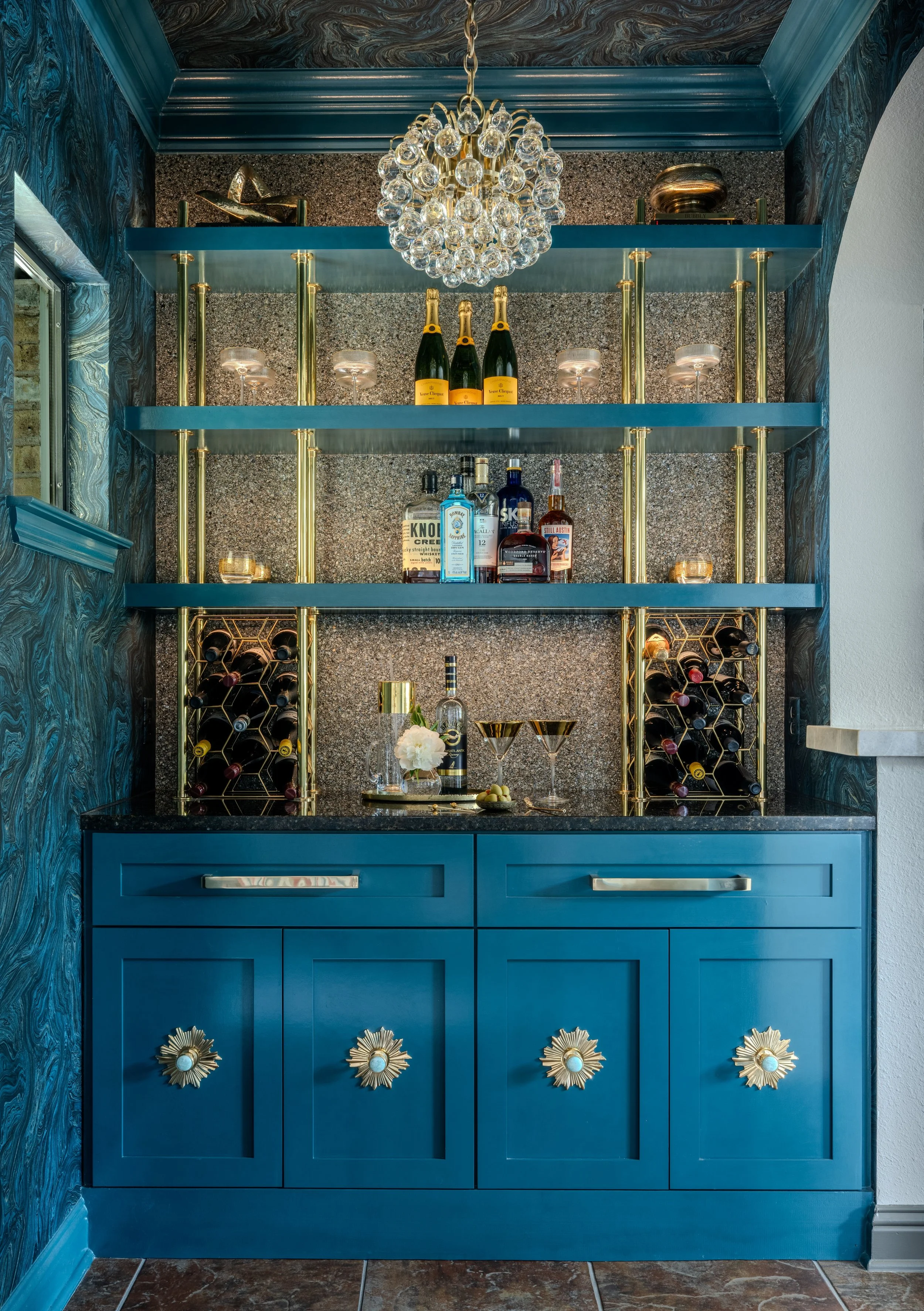 This luxury wine bar at the Schug Cove project layers dramatic blue cabinetry, brass shelving, sculptural lighting, and rich wallcovering to create a bold entertaining focal point with depth and personality. Cyndi Hopkins Design combined reflective f