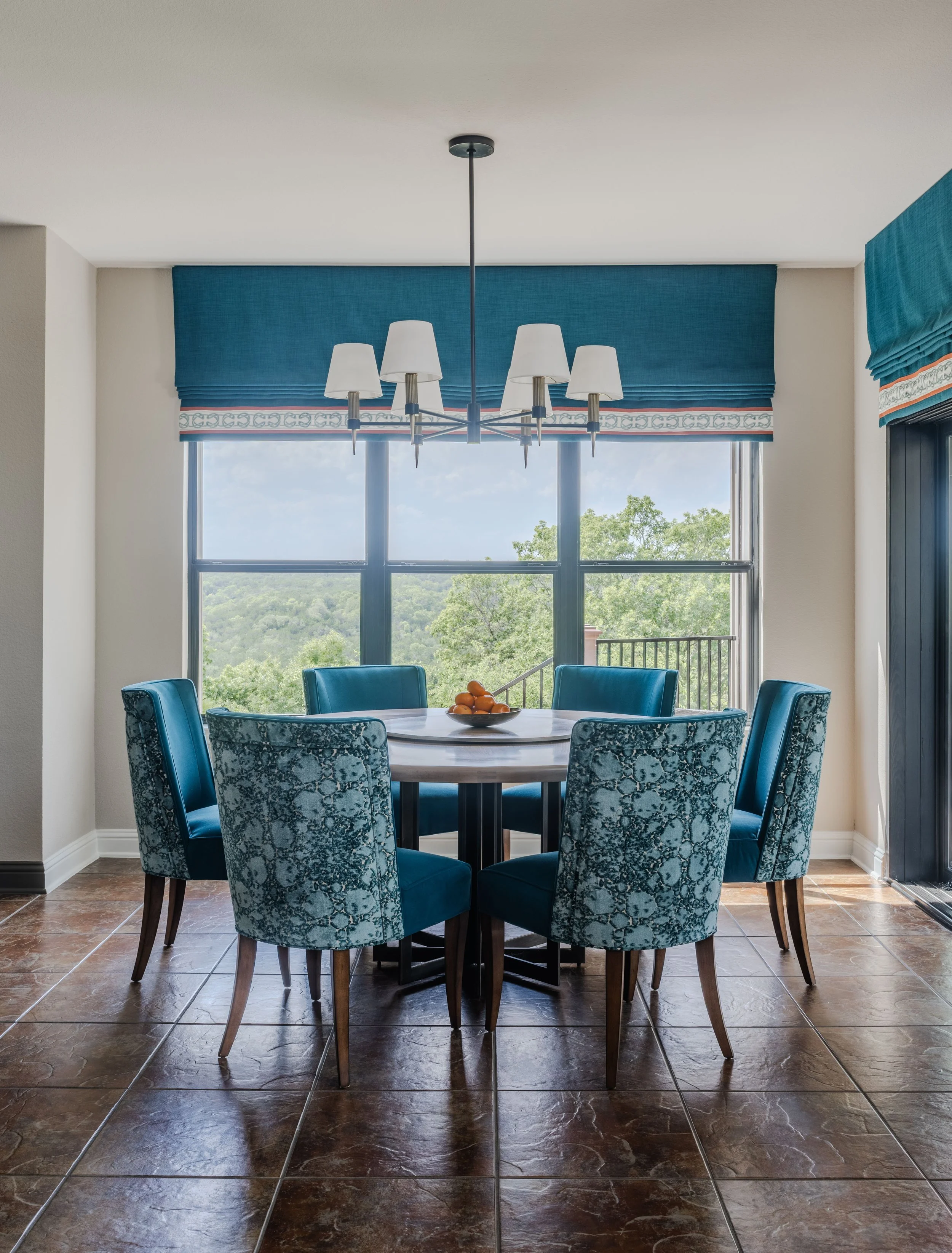 This breakfast room at the Schug Cove project was designed to feel bright, inviting, and connected to the surrounding landscape. Cyndi Hopkins Design layered vibrant blue dining chairs, custom Roman shades, and tailored lighting to create a refined e