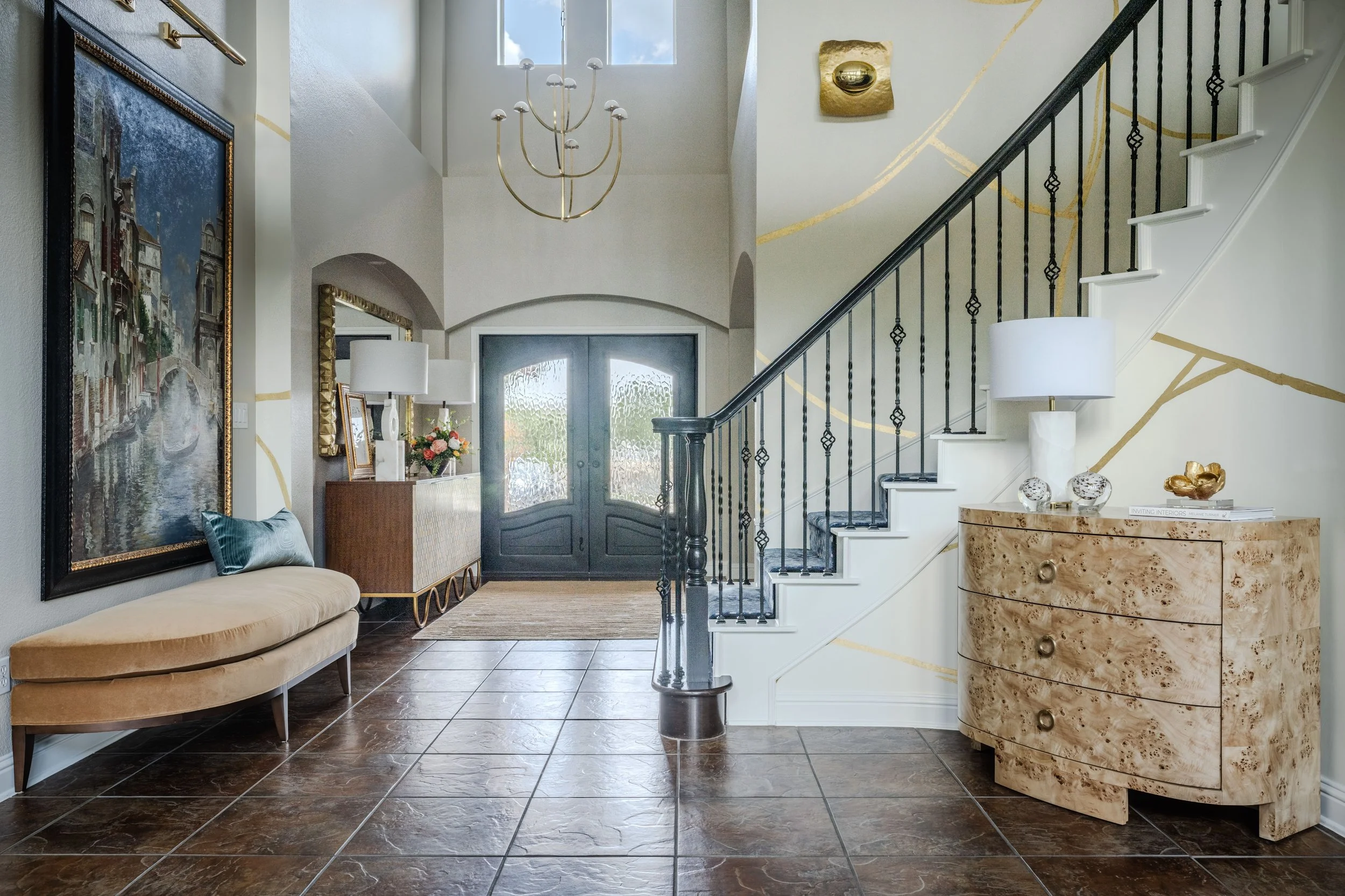 This dramatic Schug Cove foyer by Cyndi Hopkins Design creates a memorable first impression through layered textures, sculptural furnishings, and architectural detailing. The luxury entry features a curved staircase with iron balusters, a custom wall