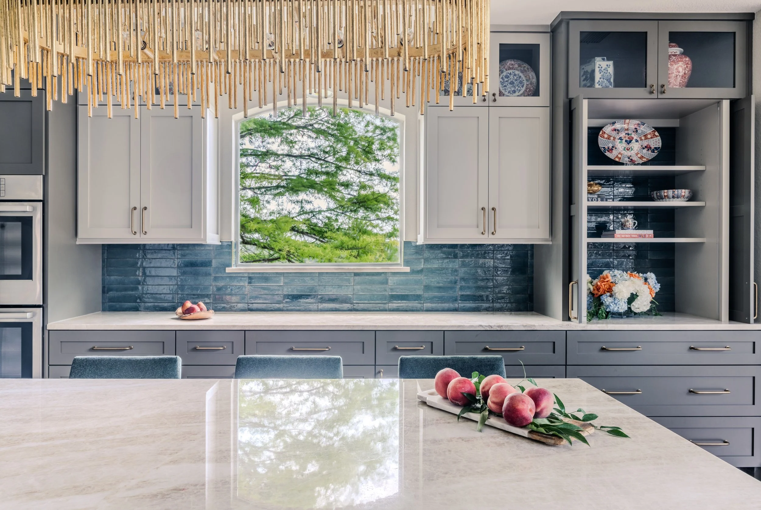This kitchen at the Schug Cove project blends custom cabinetry, layered storage, and a rich blue tile backsplash with warm quartzite surfaces to create a refined yet highly functional space. Cyndi Hopkins Design paired clean architectural lines with 