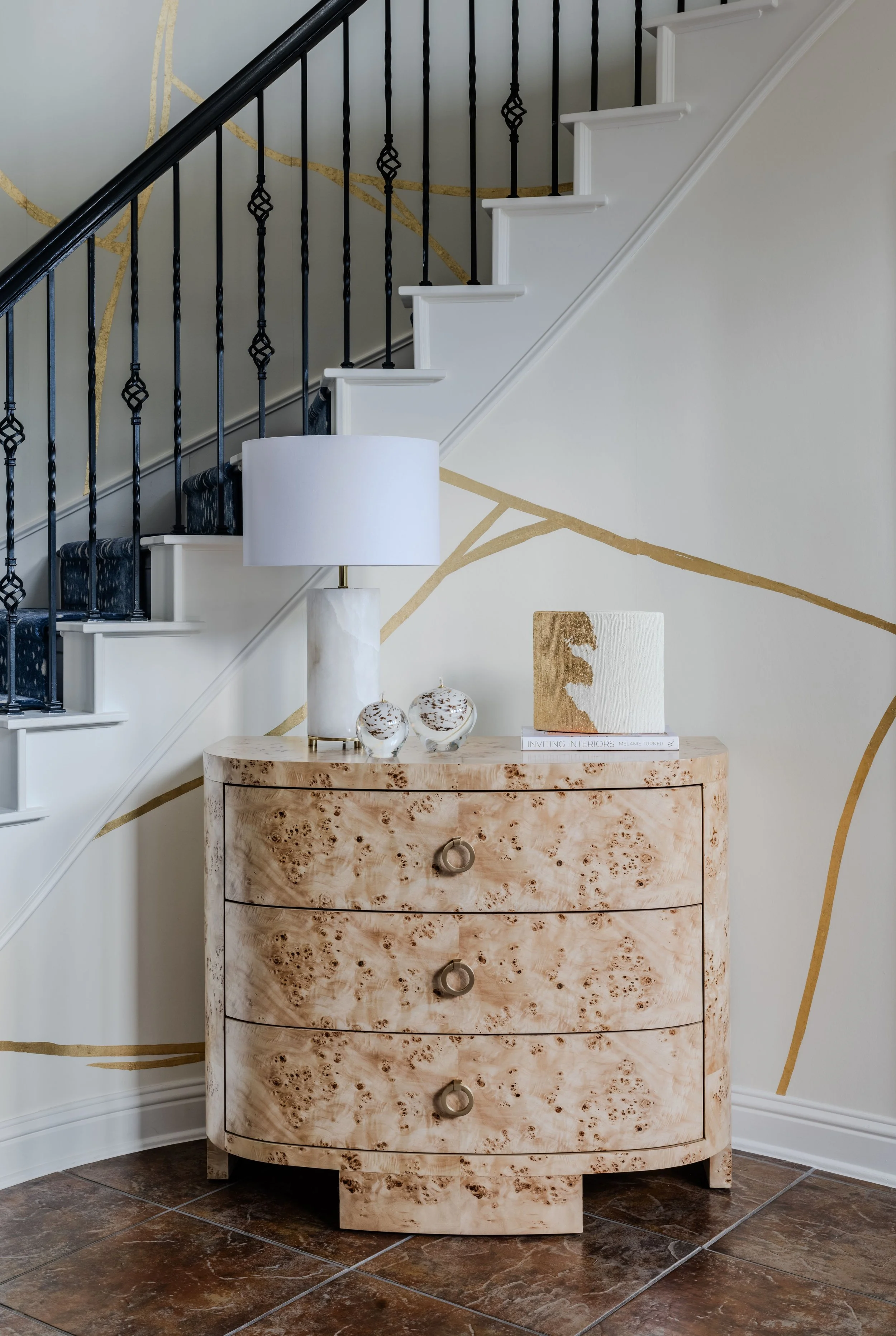 This refined foyer vignette at the Schug Cove project showcases a sculptural burl wood chest layered against a custom metallic wall mural beneath a dramatic curved staircase. Designed by Cyndi Hopkins Design, the space combines warm natural materials