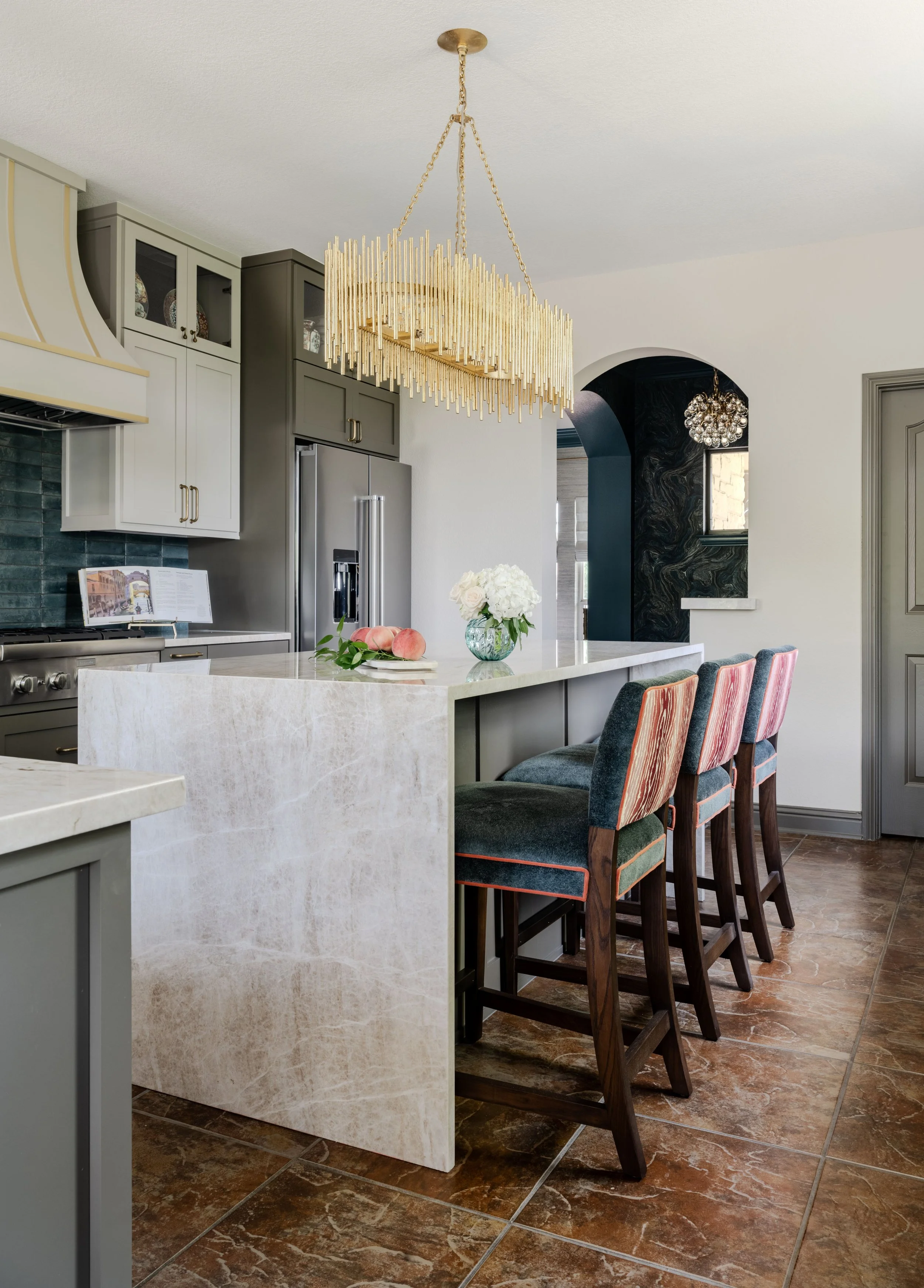 This kitchen at the Schug Cove project blends custom cabinetry, a quartzite waterfall island, and tailored barstools to create a refined gathering space designed for both entertaining and everyday living. Cyndi Hopkins Design layered sculptural light