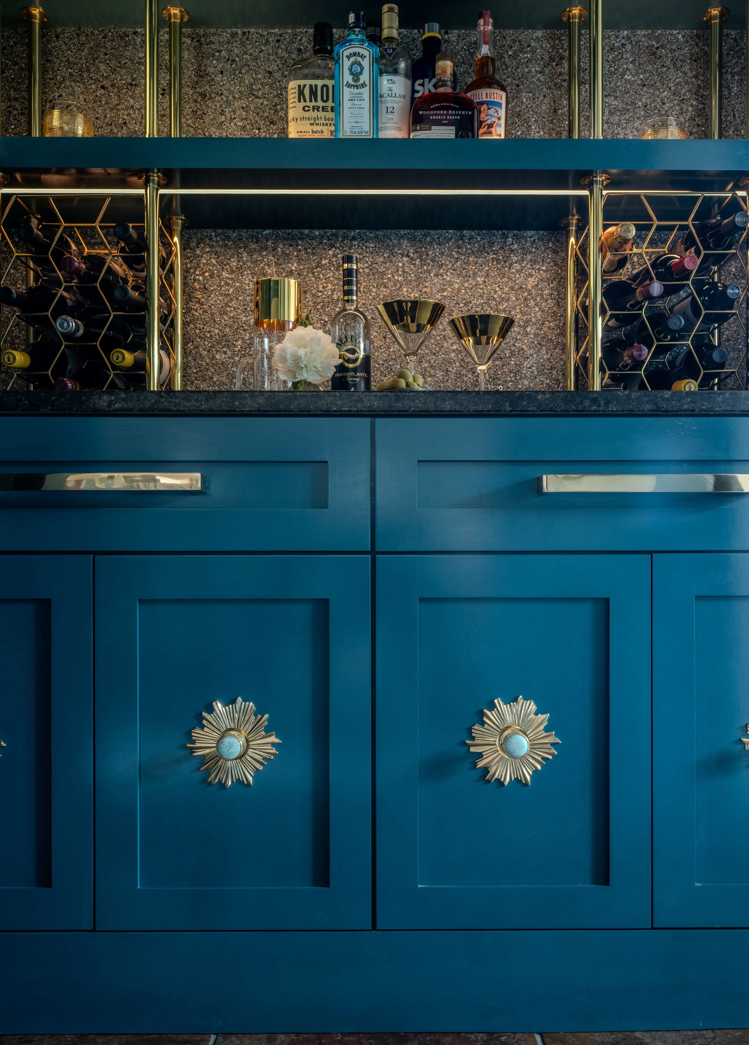 This custom wine bar at the Schug Cove project layers dramatic blue cabinetry, brass wine storage, statement hardware, and tailored lighting to create a sophisticated entertaining feature with bold personality. Cyndi Hopkins Design combined rich colo
