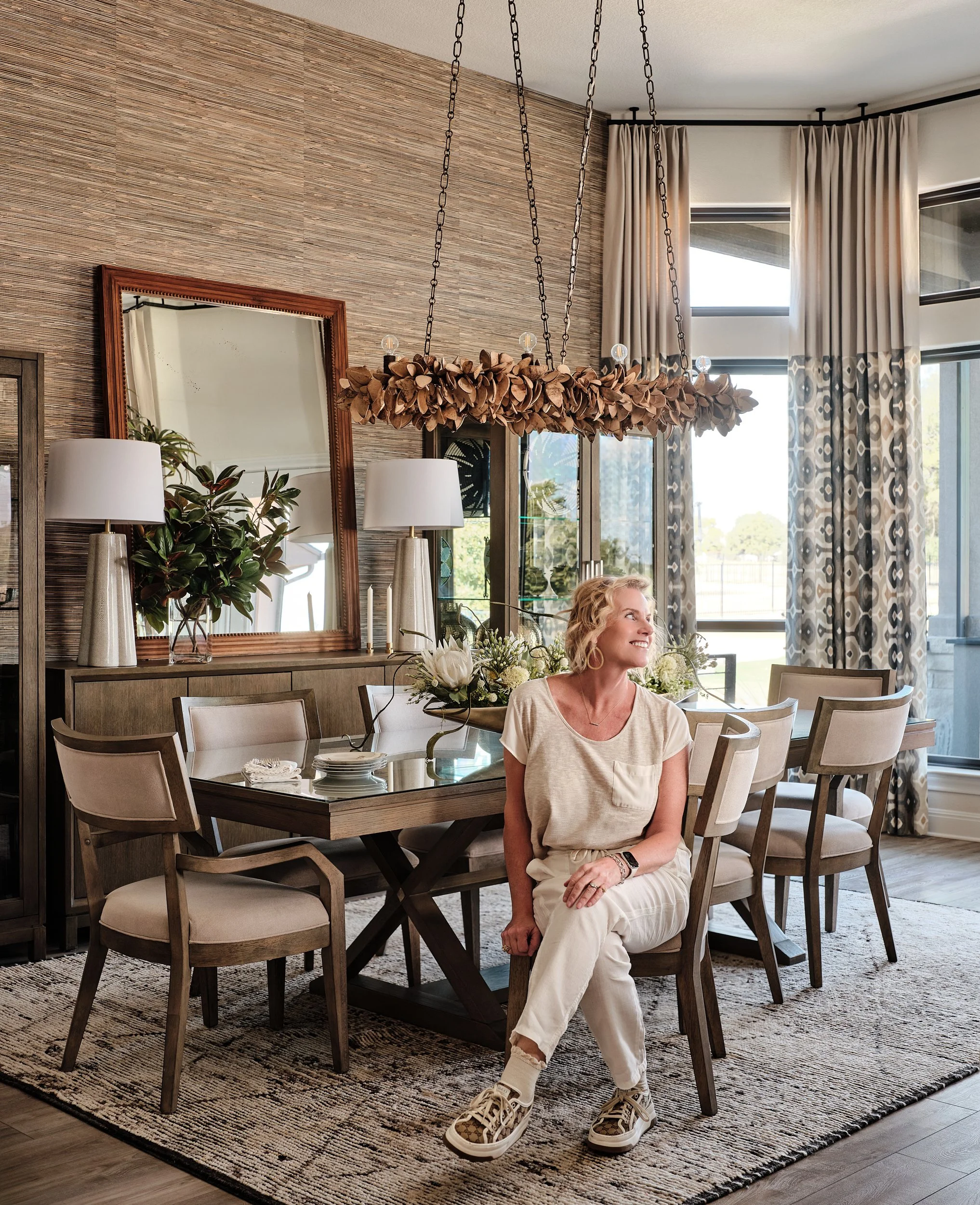 A successful interior designer sitting on a dining chair in a well-decorated dining room with a wooden table, large mirror, plants, lamps, and a chandelier, with big windows and patterned curtains.