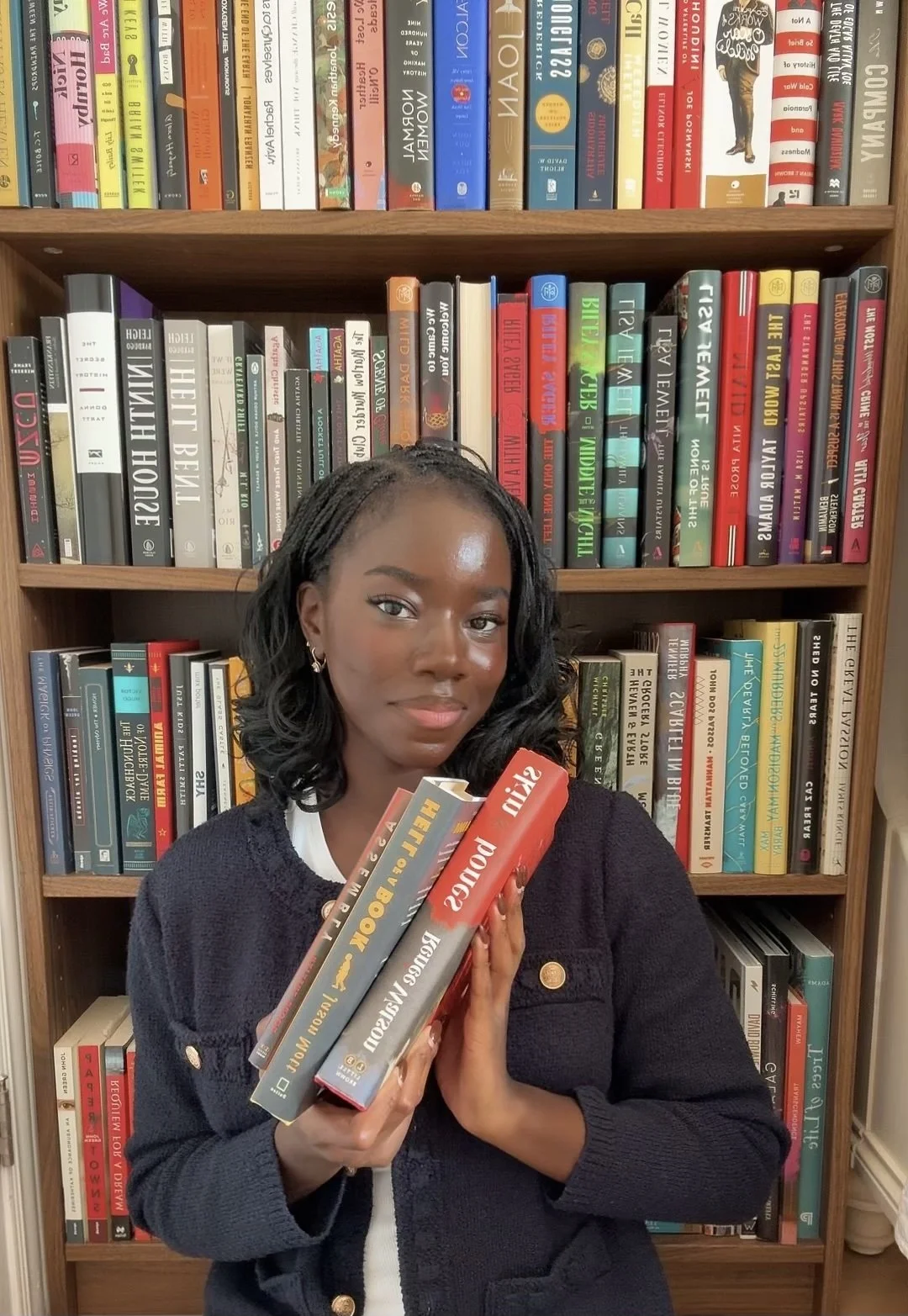 A woman holding three books stands in front of a bookshelf filled with colorful books. She has dark, shoulder-length curly hair and is wearing a black sweater. The bookshelf behind her is packed with novels and nonfiction books, with titles in various languages.