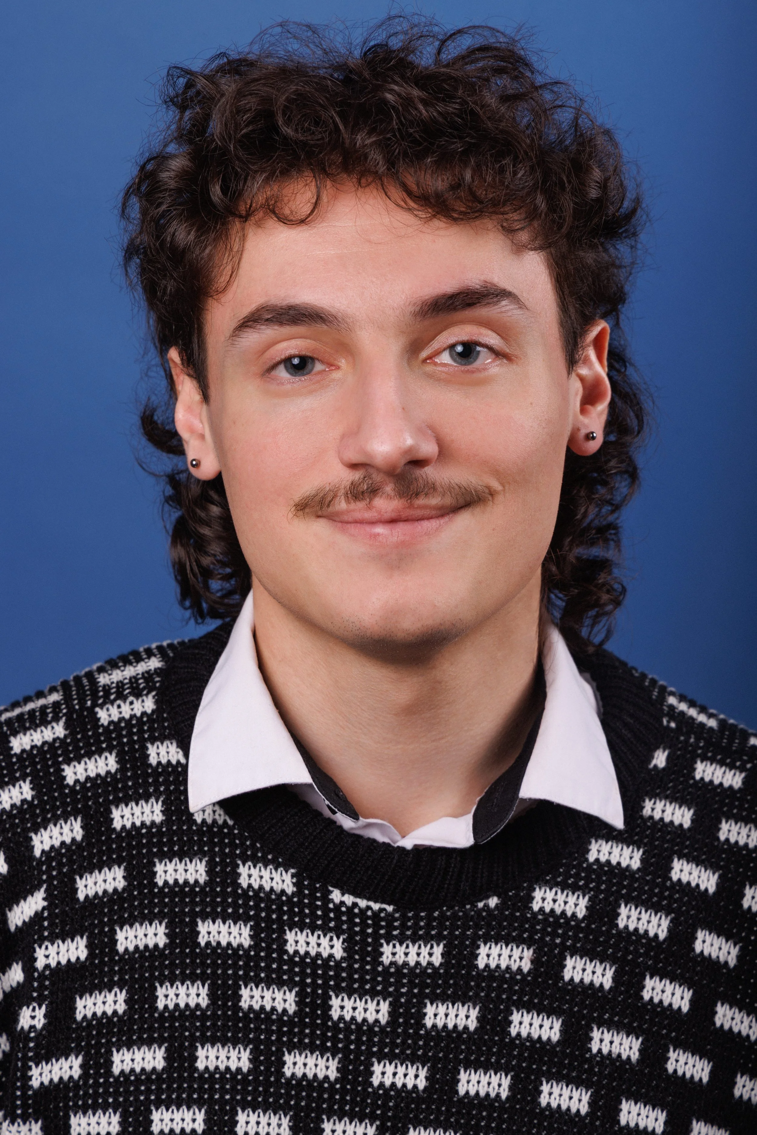 A young man with dark curly hair, blue eyes, a mustache, and earrings, smiling mildly, wearing a sweater over a collared shirt, against a blue background.
