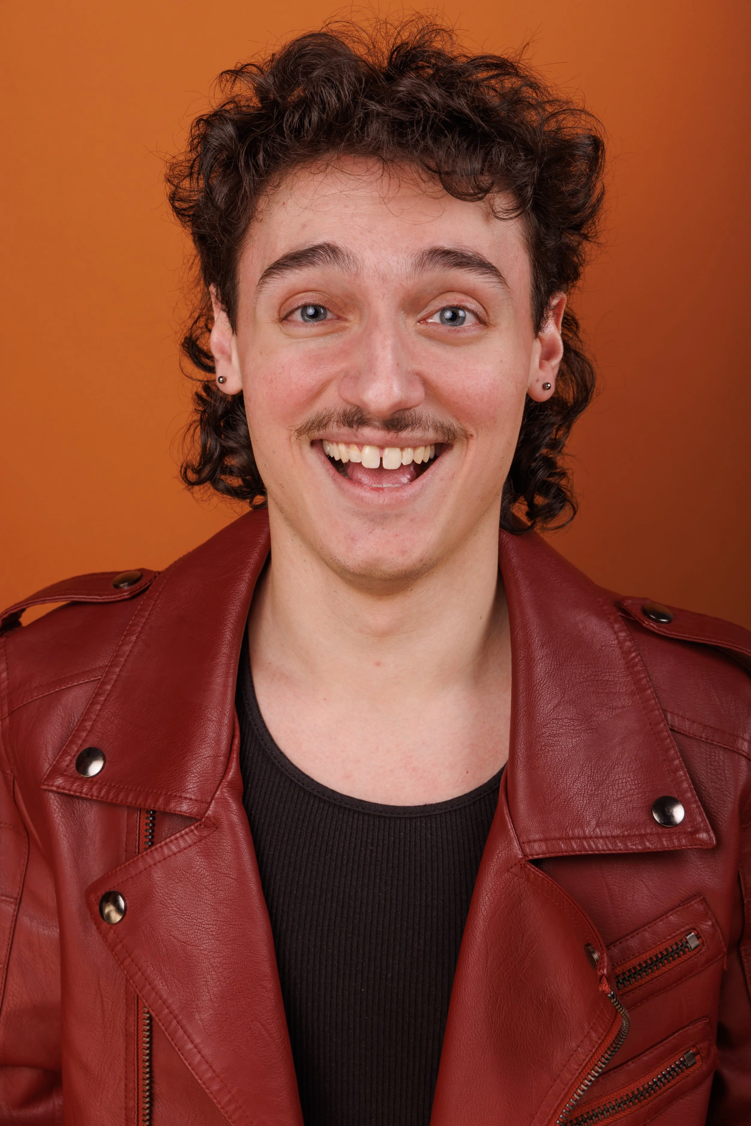 A young man with dark curly hair, light skin, blue eyes, and a mustache, smiling openly, wearing a red leather jacket over a black shirt, against an orange background.