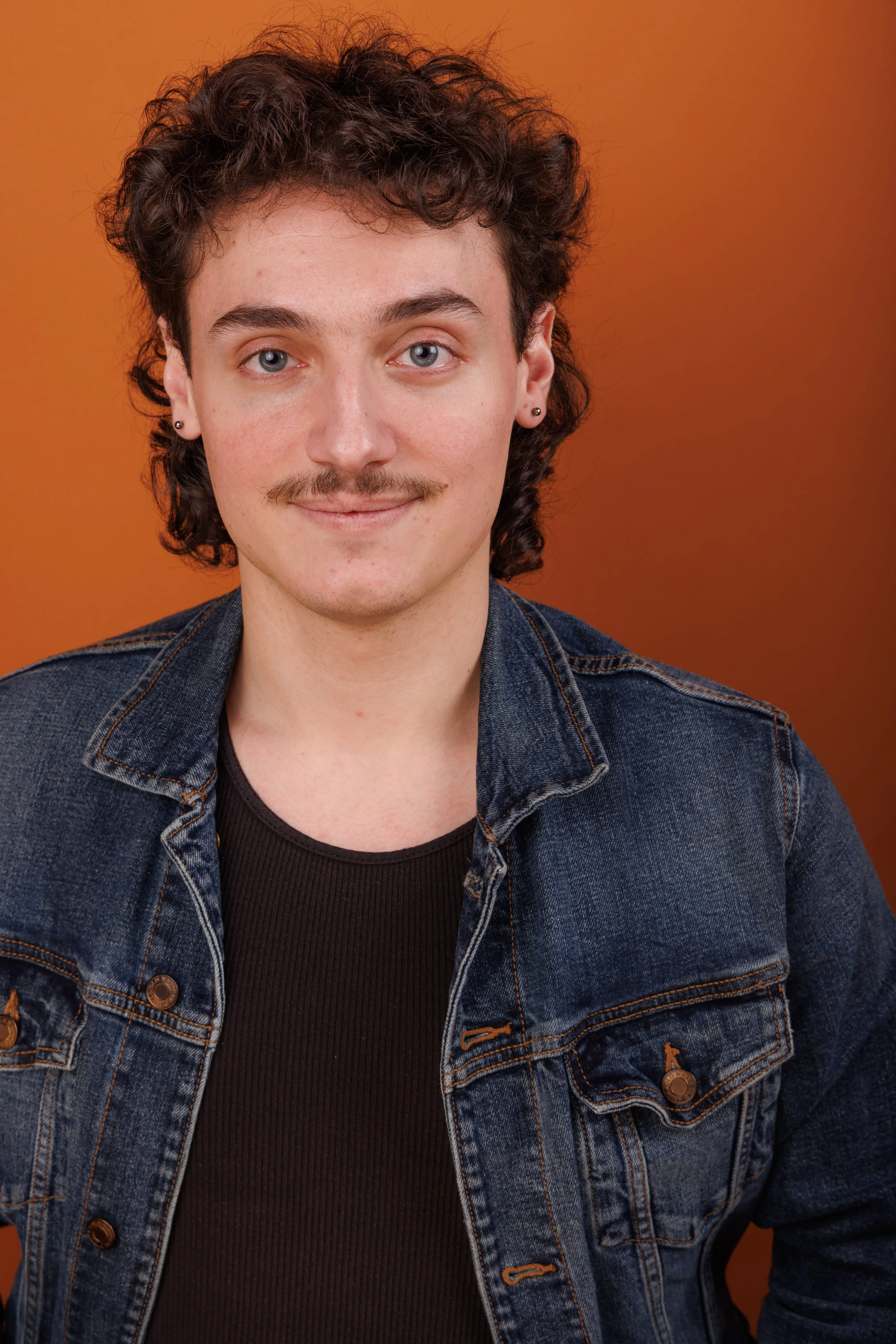 Portrait of a young man with curly hair, mustache, and earrings, wearing a denim jacket and black shirt, standing against an orange background.
