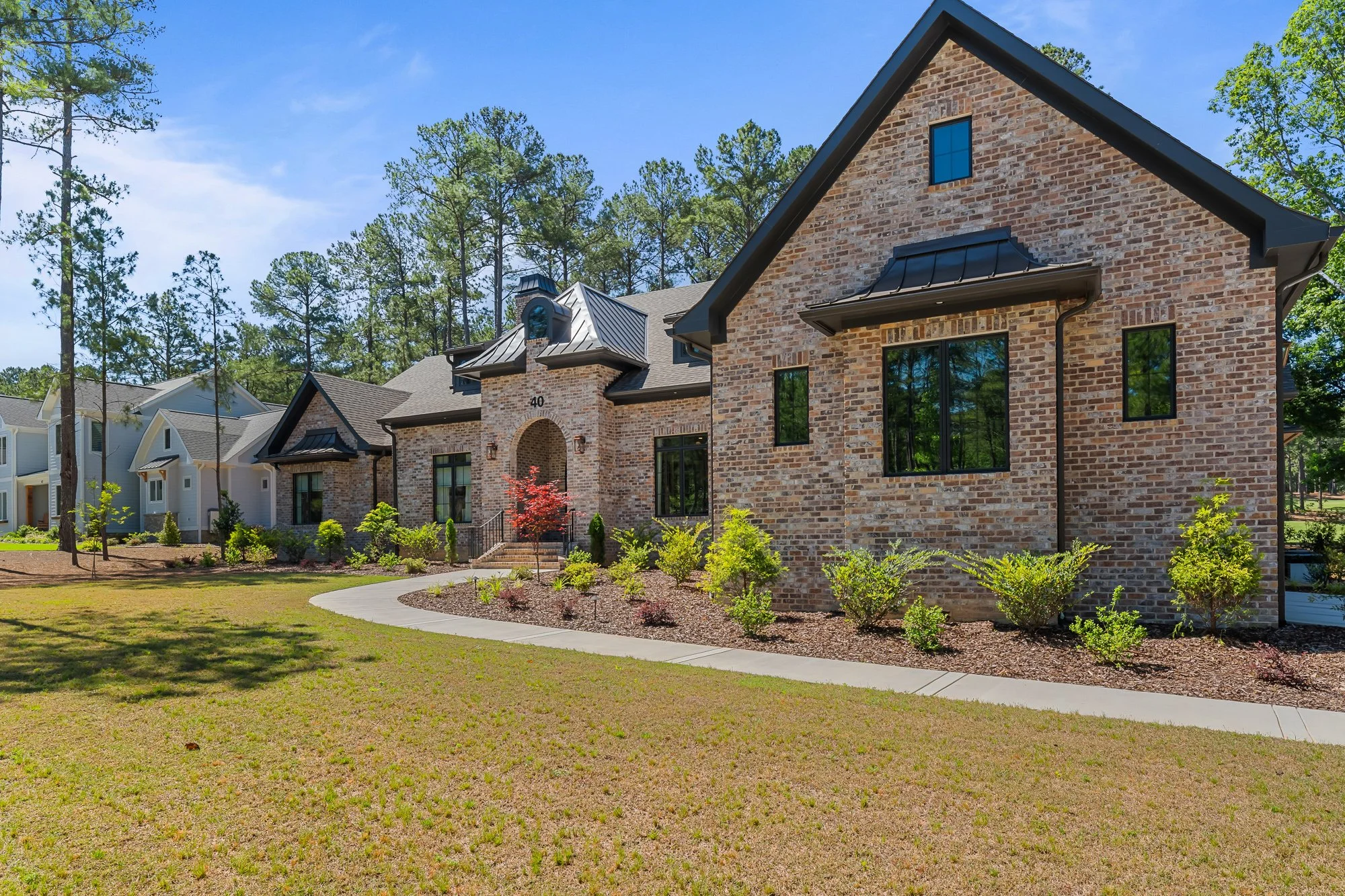Front three-quarter view of a luxury European-inspired custom brick home by FRS Homes featuring steep gables, black metal accents, arched entryway, and professionally landscaped grounds in Southern Pines, North Carolina.