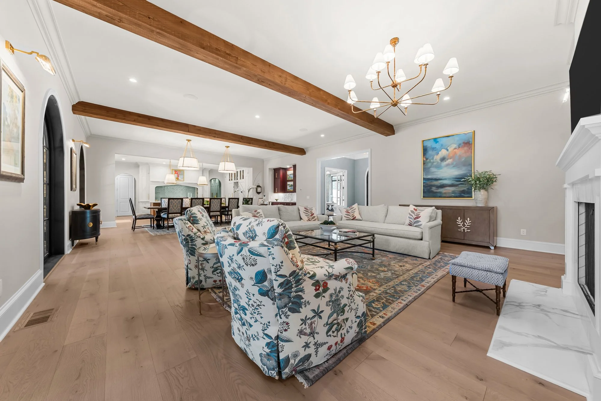 Open-concept luxury great room by FRS Homes featuring exposed wood ceiling beams, marble fireplace, designer chandelier, hardwood floors, and seamless flow into the dining room and chef's kitchen.