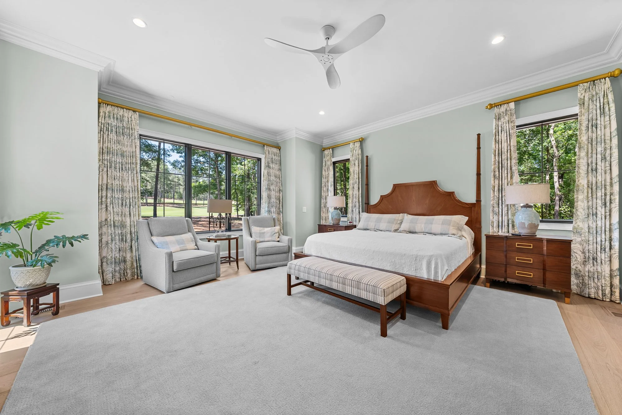 Luxury primary bedroom by FRS Homes featuring oversized picture windows, custom wood bed, hardwood floors, designer drapery, and a spacious sitting area overlooking the wooded landscape.