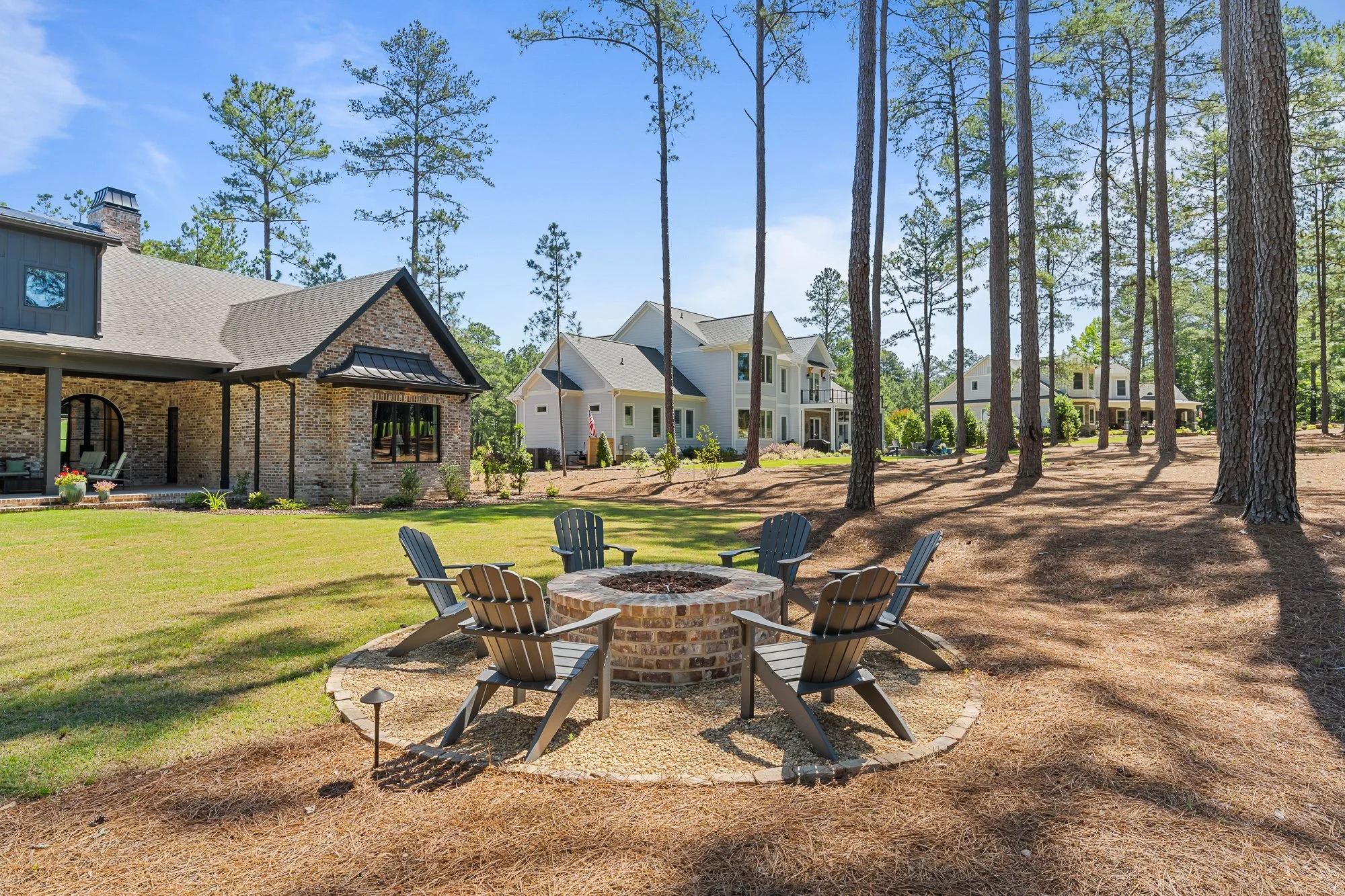 Custom backyard by FRS Homes featuring a circular brick fire pit, Adirondack seating, mature pine trees, and a covered outdoor living area designed for entertaining and relaxing.