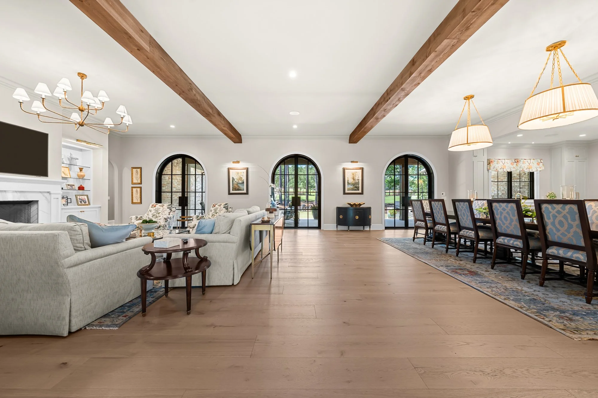 Luxury open-concept living and dining room by FRS Homes featuring exposed wood ceiling beams, arched steel-framed doors, designer lighting, wide-plank hardwood floors, and seamless indoor-outdoor living.