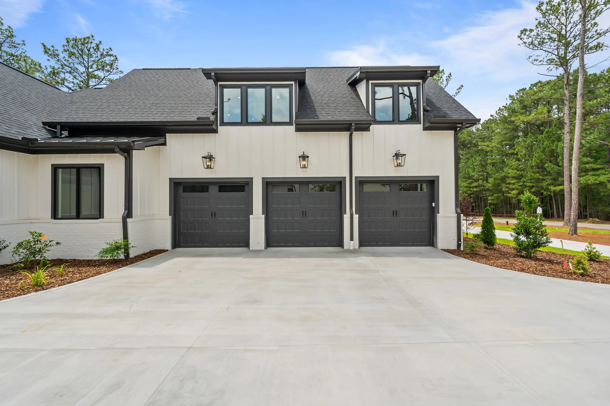 Three-car garage at Foxglove Run featuring dark garage doors, white vertical siding, black trim, dormer windows, and a spacious concrete driveway.