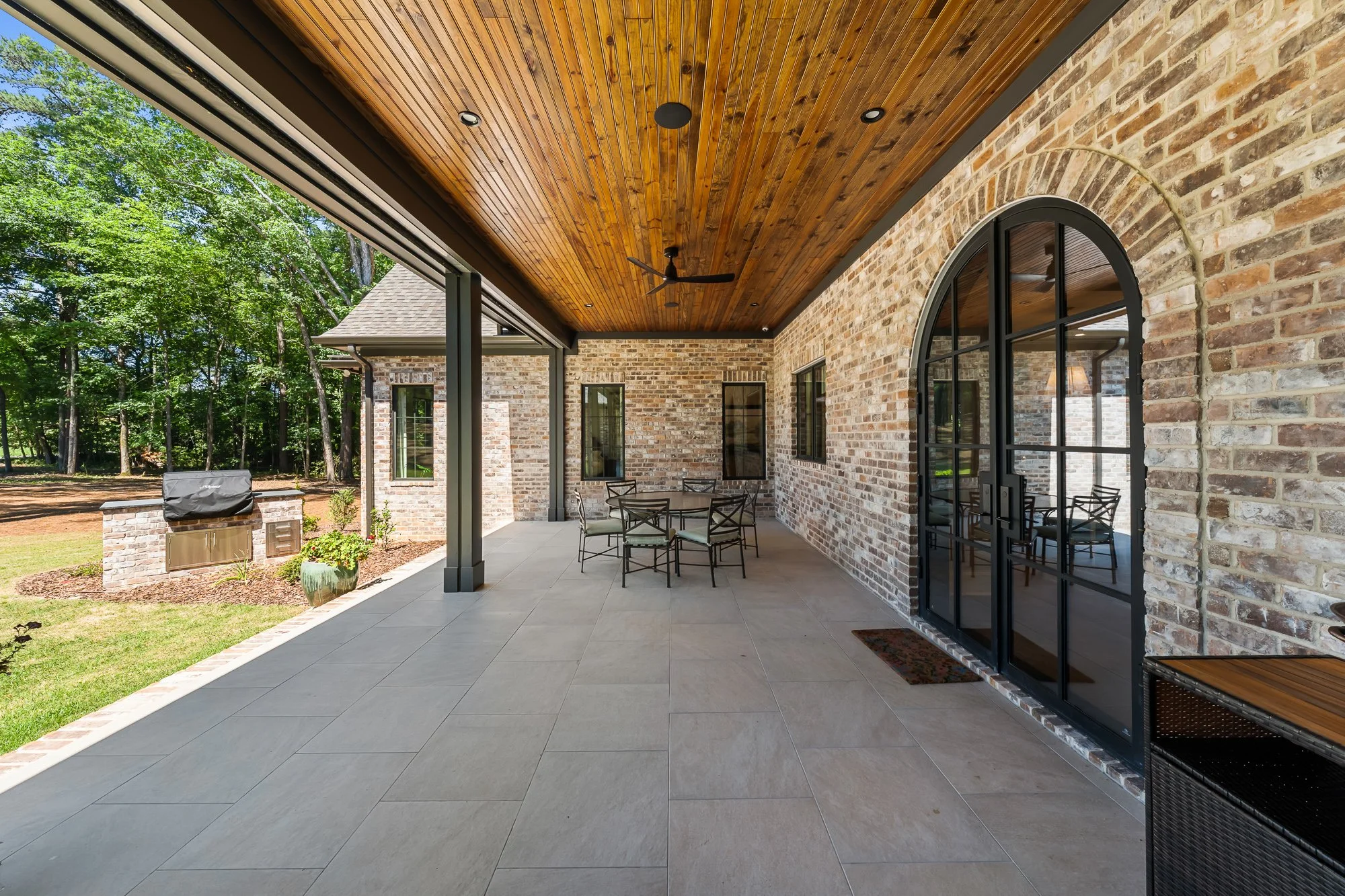 Covered outdoor living patio by FRS Homes featuring arched black steel doors, stained wood ceiling, built-in grilling station, and spacious entertaining area overlooking the landscaped backyard.