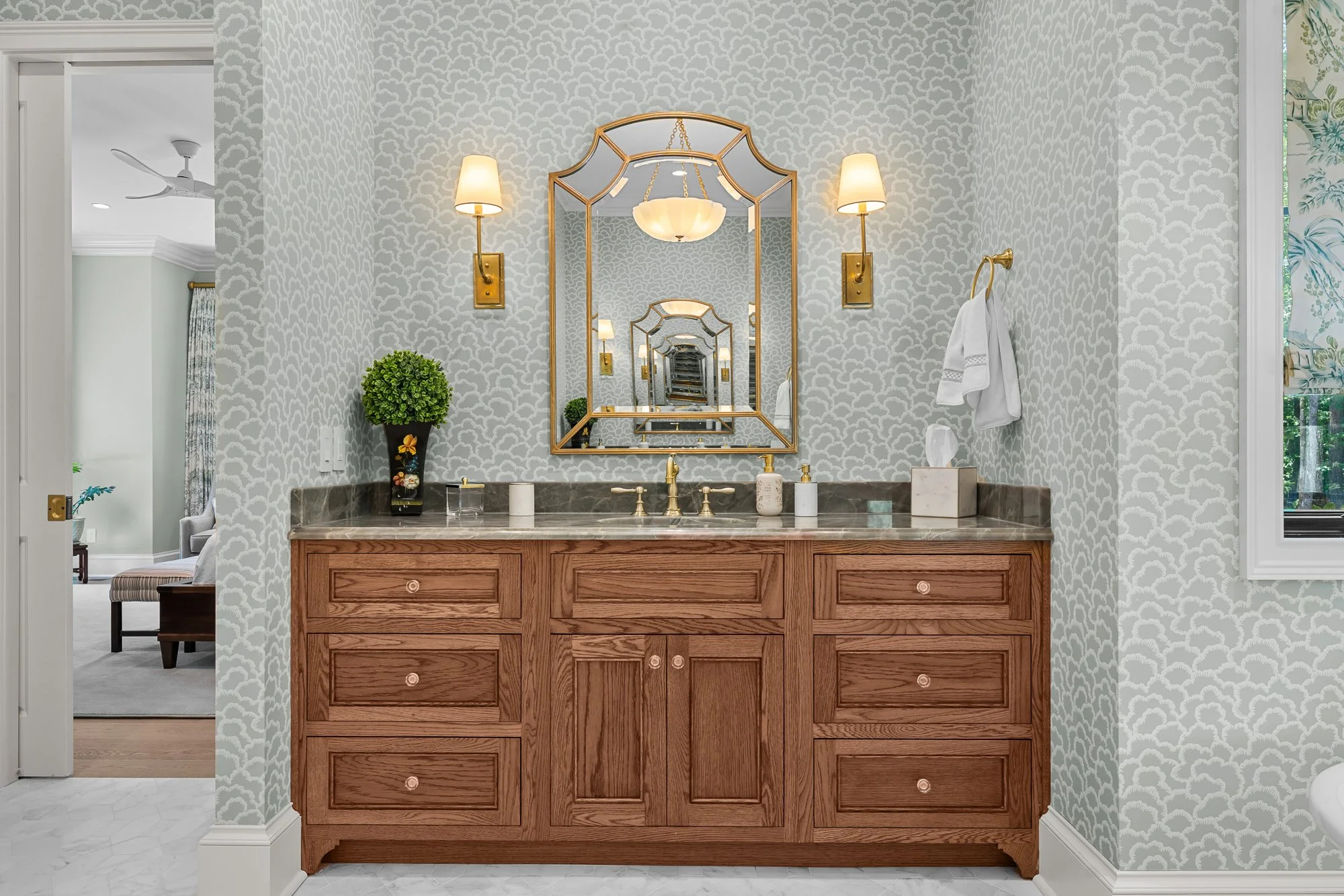 Luxury primary bathroom vanity by FRS Homes featuring custom oak cabinetry, marble countertops, designer brass fixtures, patterned wallpaper, and elegant wall sconces.