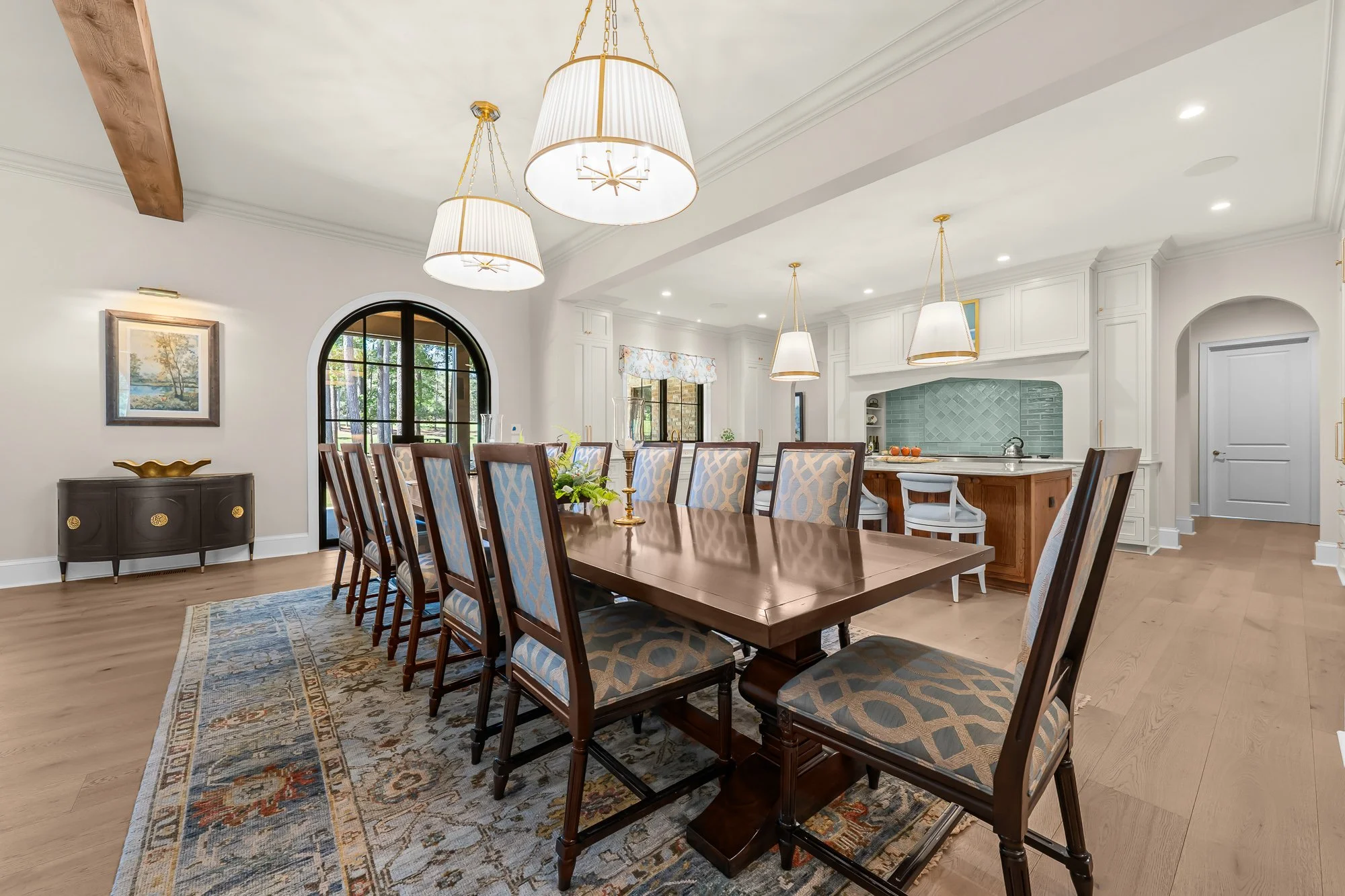 Open-concept luxury dining room and chef's kitchen by FRS Homes featuring oversized designer pendant lighting, exposed wood beam, custom cabinetry, large island, and arched steel patio doors.