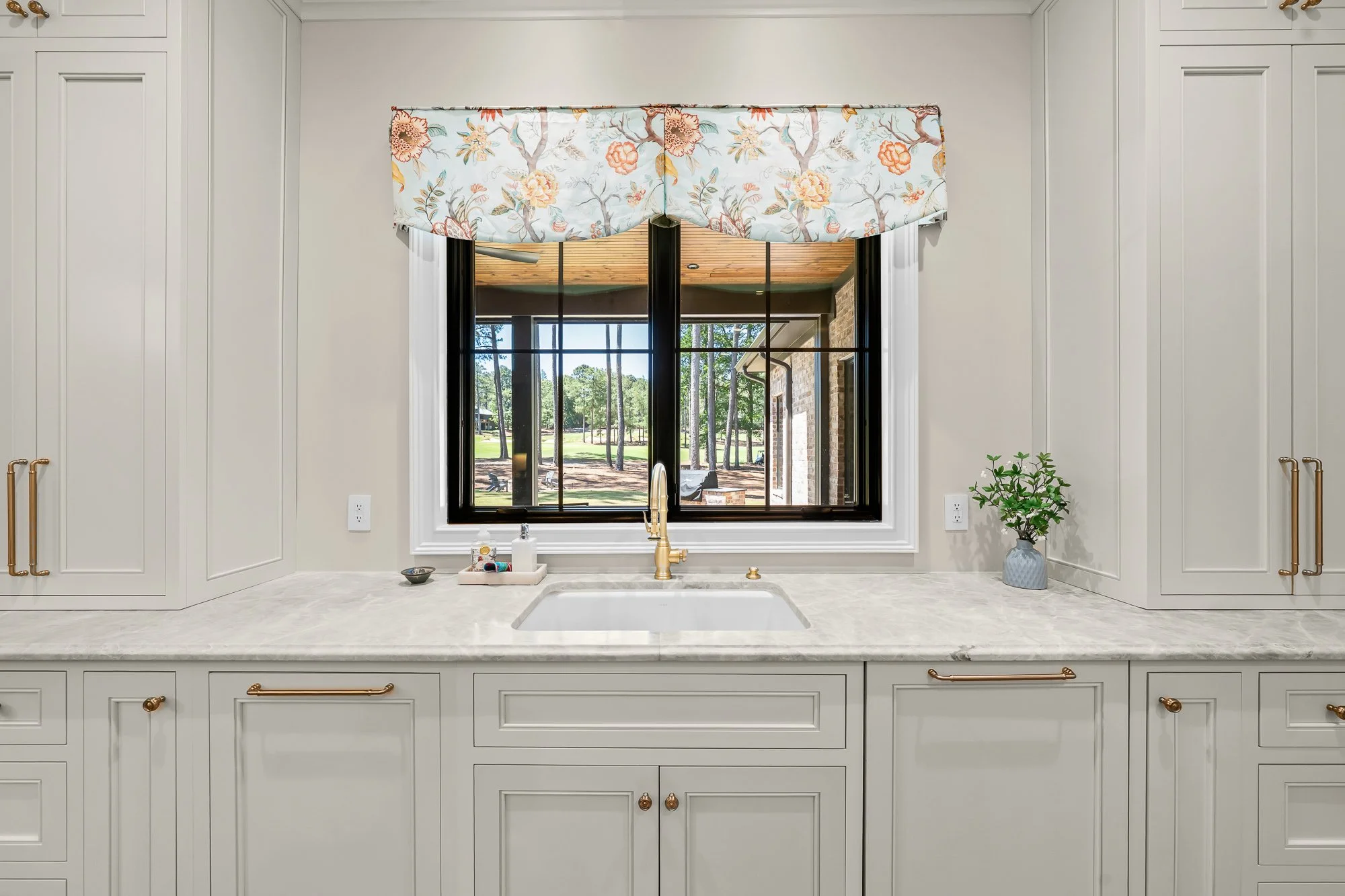 Custom chef's kitchen sink by FRS Homes featuring marble countertops, custom white cabinetry, brushed brass hardware, and a black-framed picture window overlooking the covered outdoor living space.