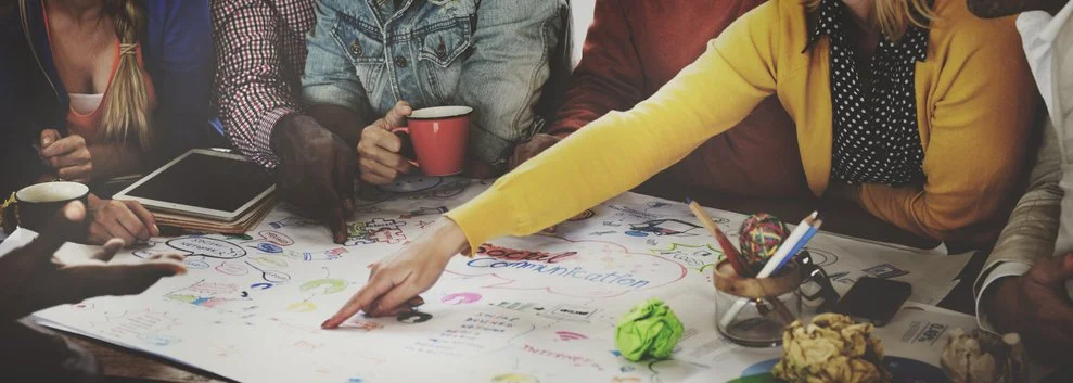 People gathered around a table working on a large sheet of paper with colorful notes and drawings, some holding mugs, in a collaborative setting.