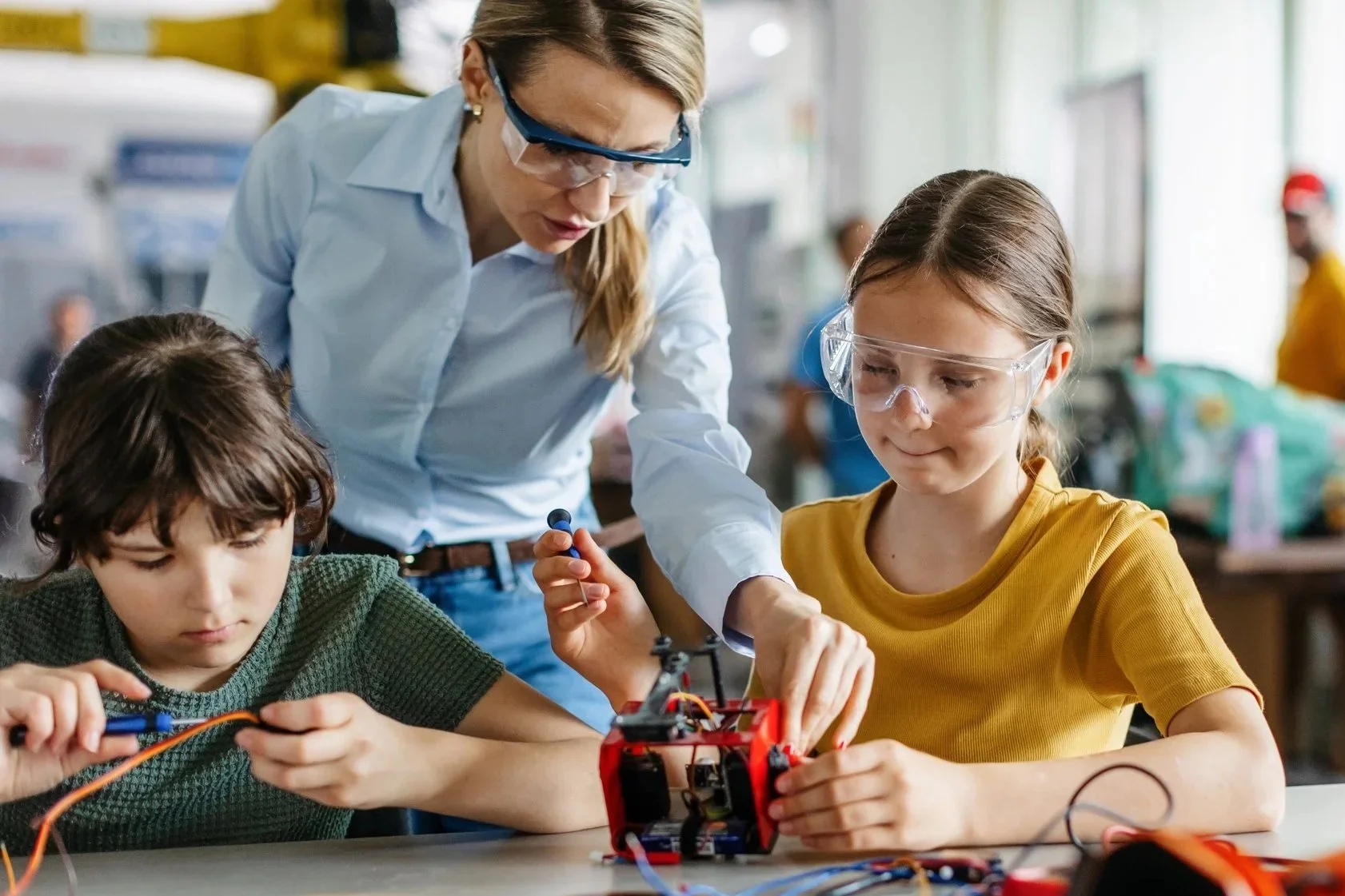 A woman instructs two children working on a small robot at a table in a classroom or workshop.