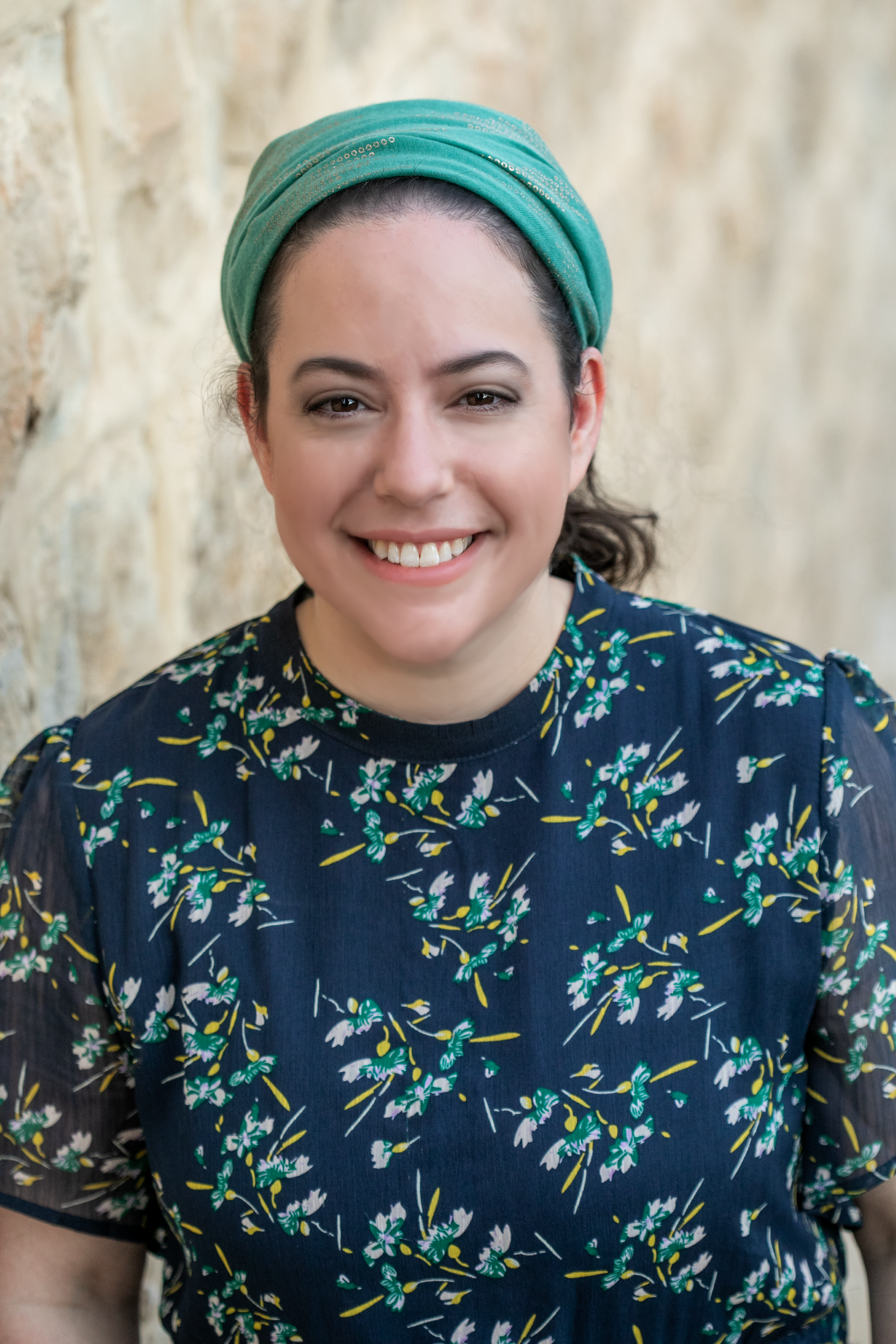 A woman with a green headwrap smiling, wearing a dark floral dress, standing against a textured wall.