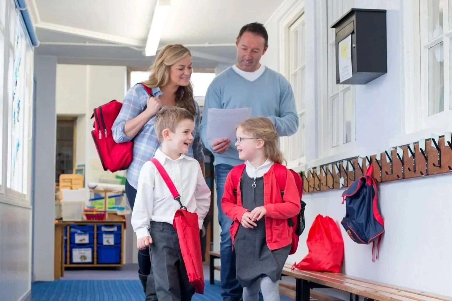 A school hallway with two young children, a boy and a girl, talking and smiling, with a woman and a man, possibly teachers or parents, standing nearby. The children are with backpacks, and the hallway has cubbies and hooks.