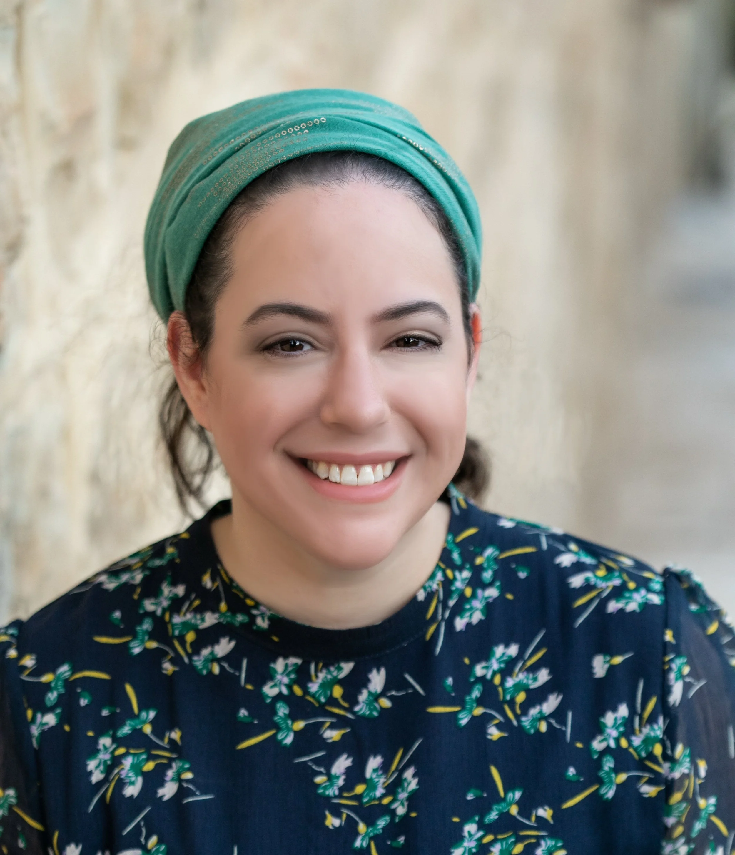 A woman smiling, wearing a turquoise headscarf and a dark floral blouse.