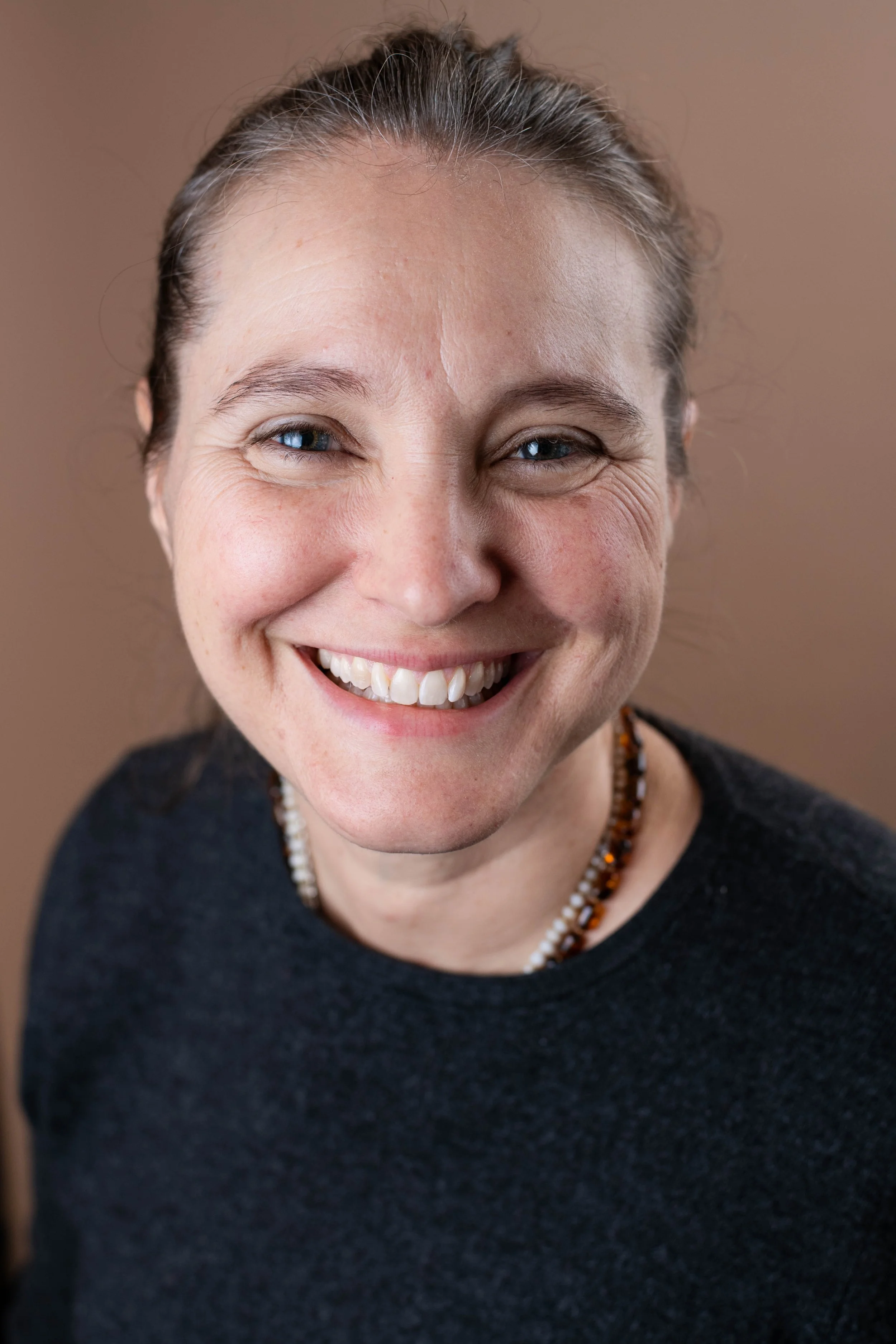 Close-up of a woman smiling with blue eyes, dark hair tied back, wearing a dark sweater and beaded necklace, against a brown background.