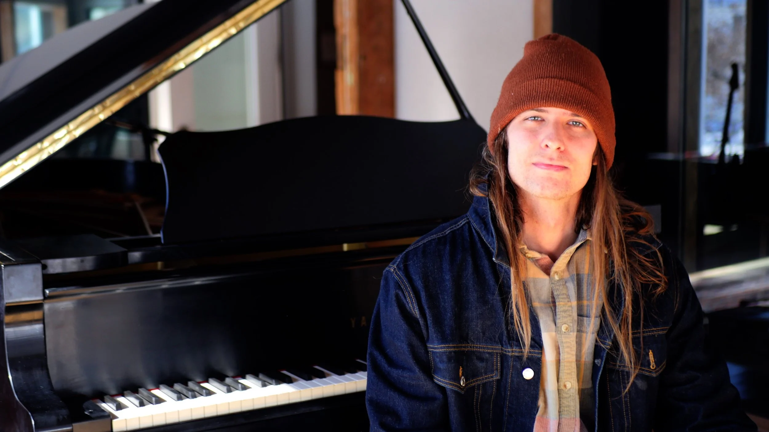 A young man with long hair wearing a brown beanie, denim jacket, and a plaid shirt sitting next to a grand piano.