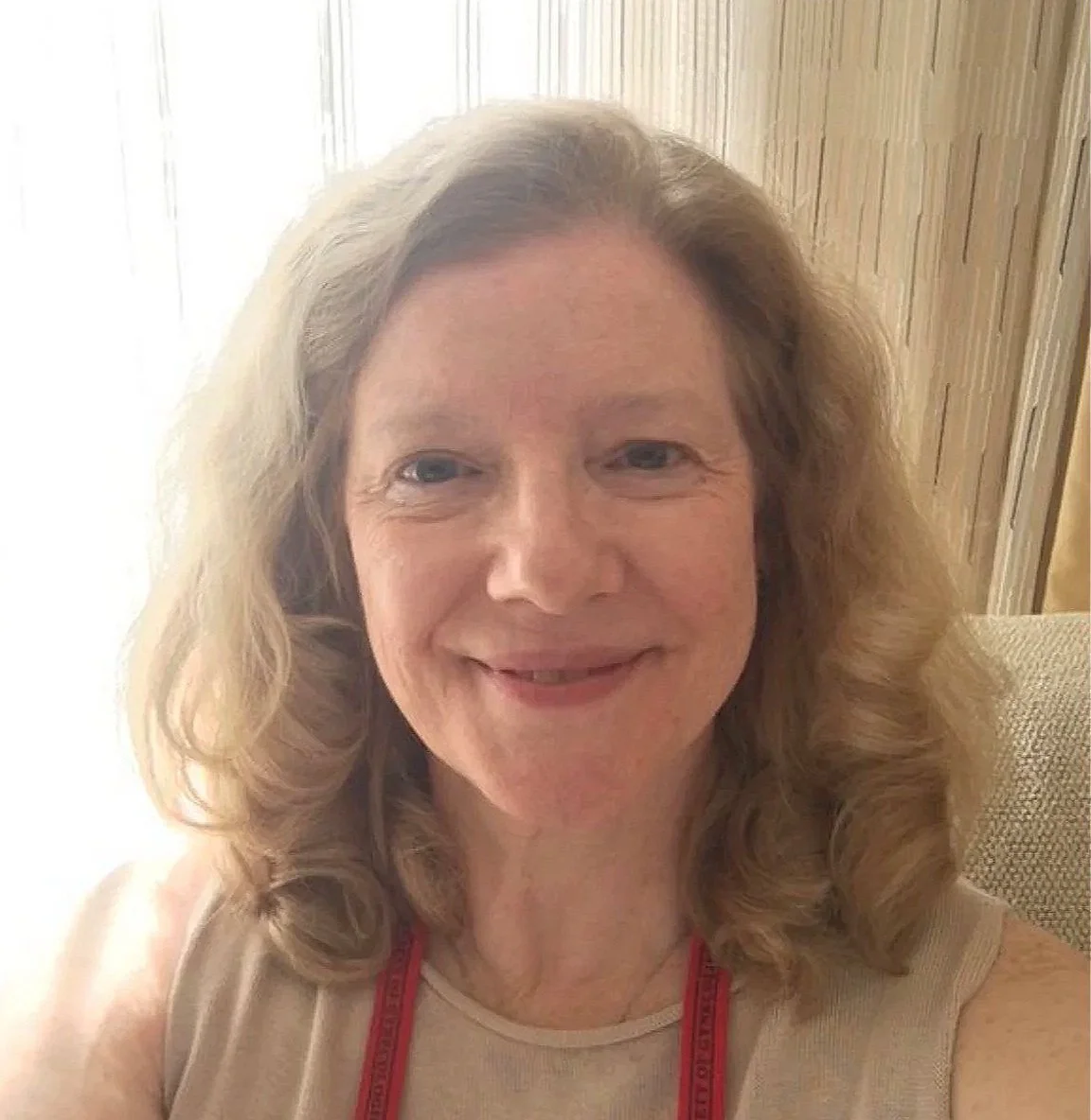 A woman with long, wavy blonde hair smiling, wearing a beige sleeveless top, sitting indoors near a window with wooden paneling in the background.