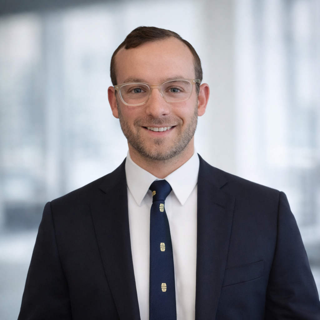 A man with short brown hair, glasses, and a beard, smiling, wearing a navy suit, white shirt, and a navy tie with small emblem, standing in front of a blurred window background.