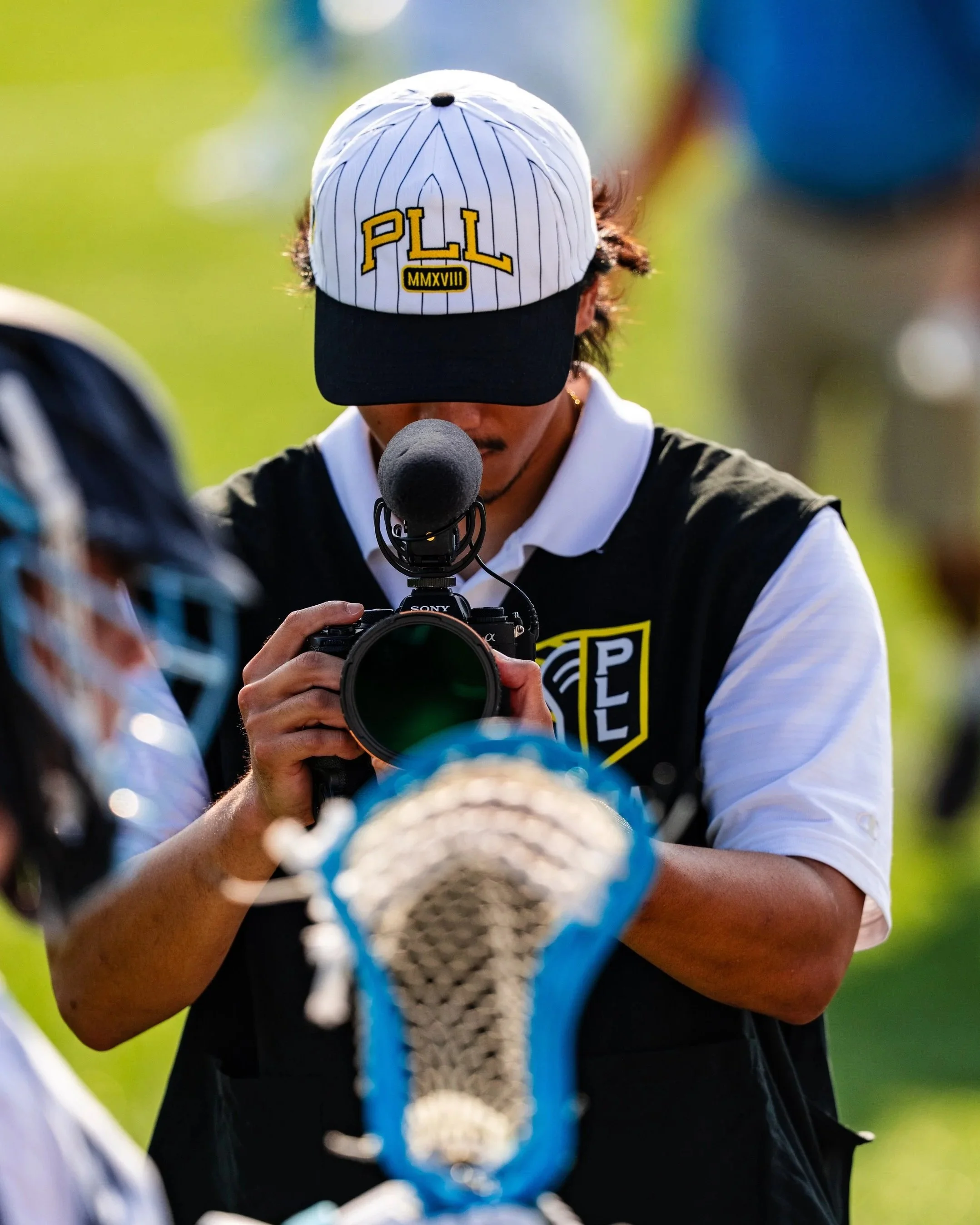 Person taking a photograph with a camera during a lacrosse game, wearing a baseball cap with 'PLL' and Roman numerals.