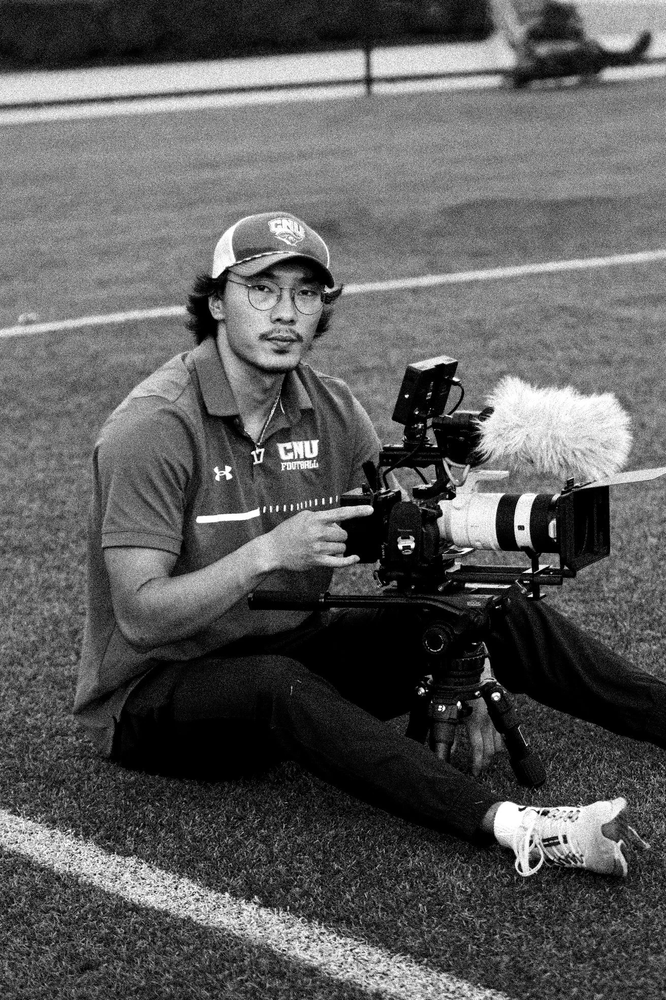 A young man wearing glasses, a baseball cap, and a sports shirt, sitting on the ground at a football field, operating a professional camera mounted on a tripod.