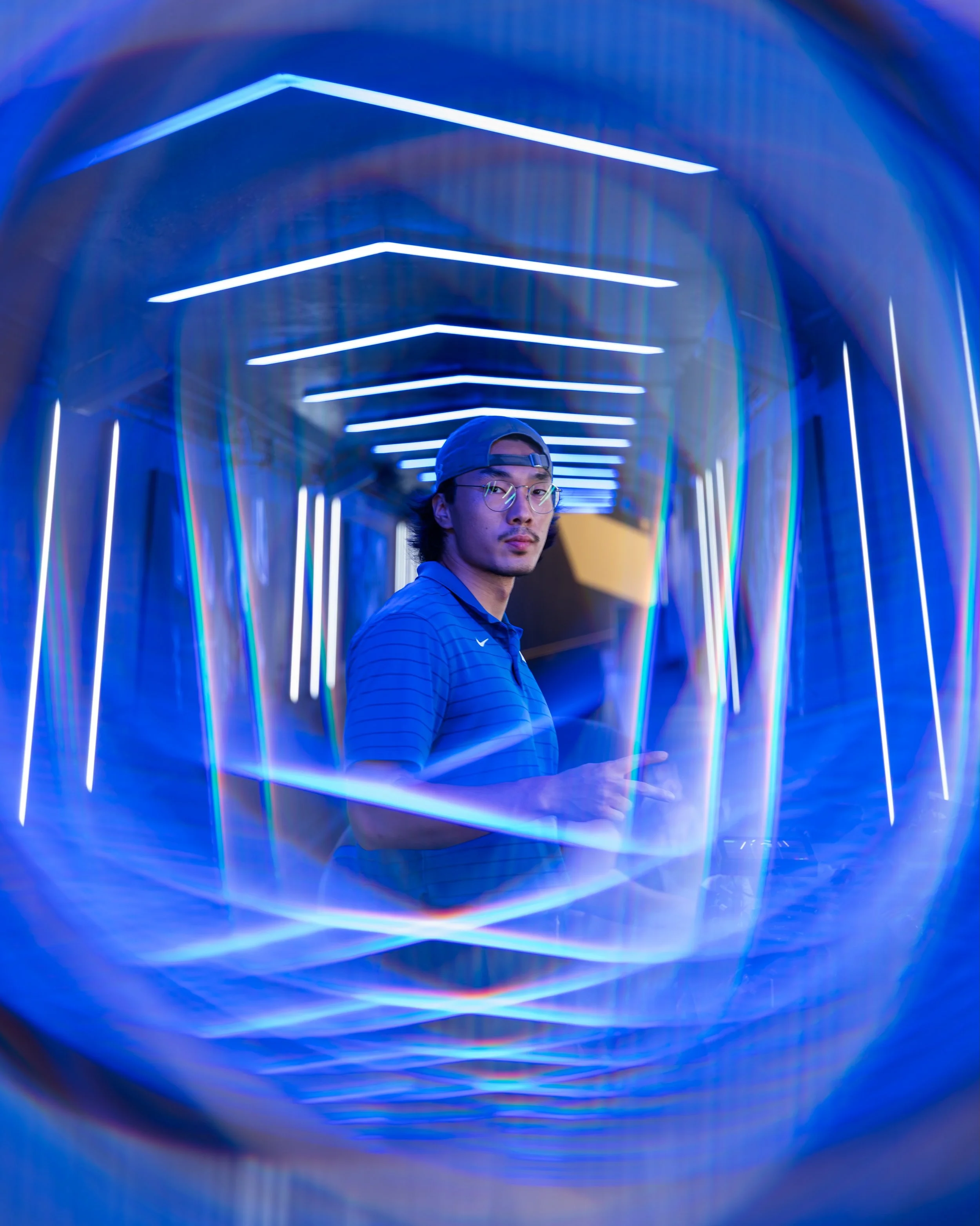 A person wearing a baseball cap and glasses standing in a tunnel with vibrant blue and purple neon lights creating a geometric pattern.