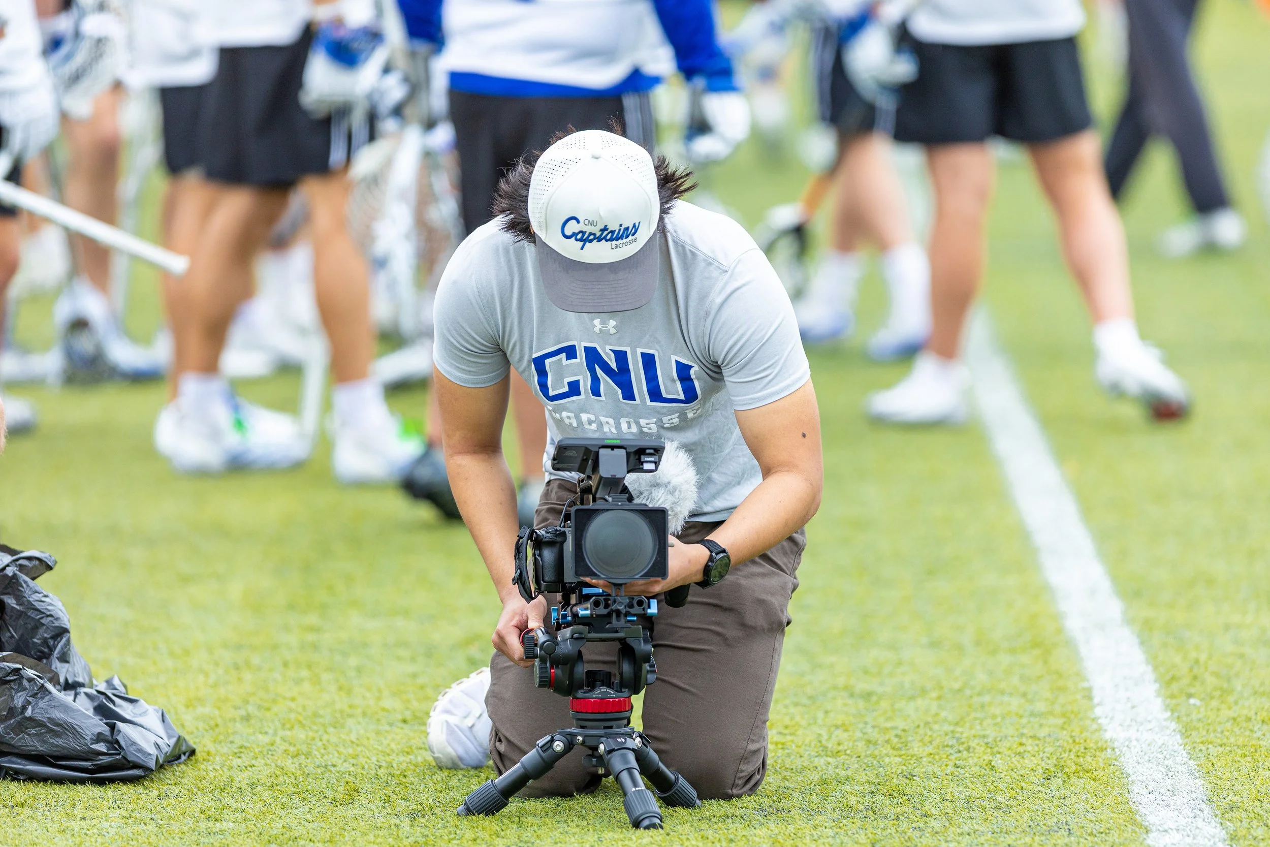 A person kneeling on grass with a video camera, wearing a gray baseball cap and a light gray T-shirt with 'CNU Lacrosse' printed on it, capturing footage during a sporting event with players in the background.