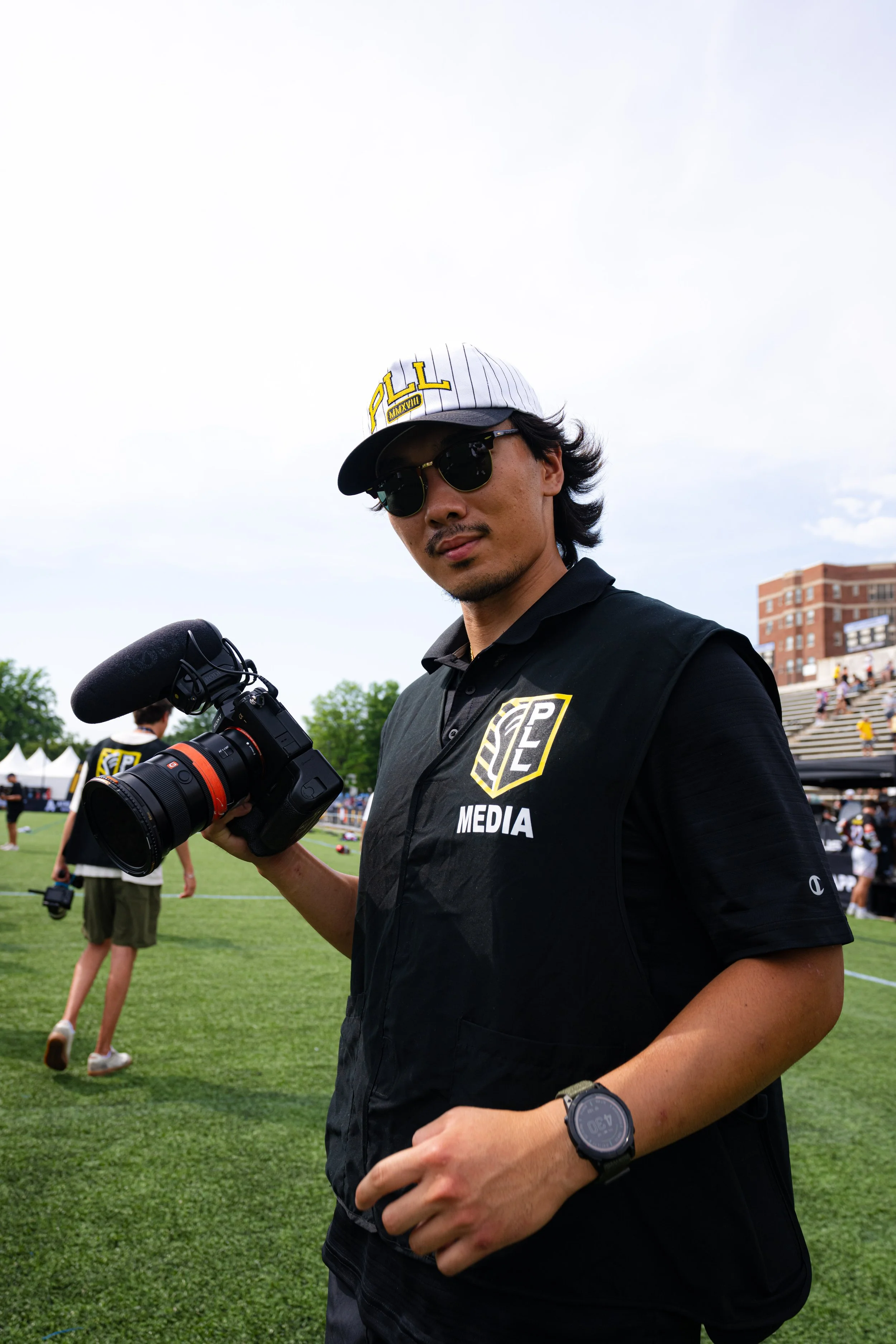 A man wearing sunglasses, a baseball cap with 'ALL' on it, a black shirt with 'MEDIA' on it, and a wristwatch, holding a professional camera with a microphone during an outdoor event.