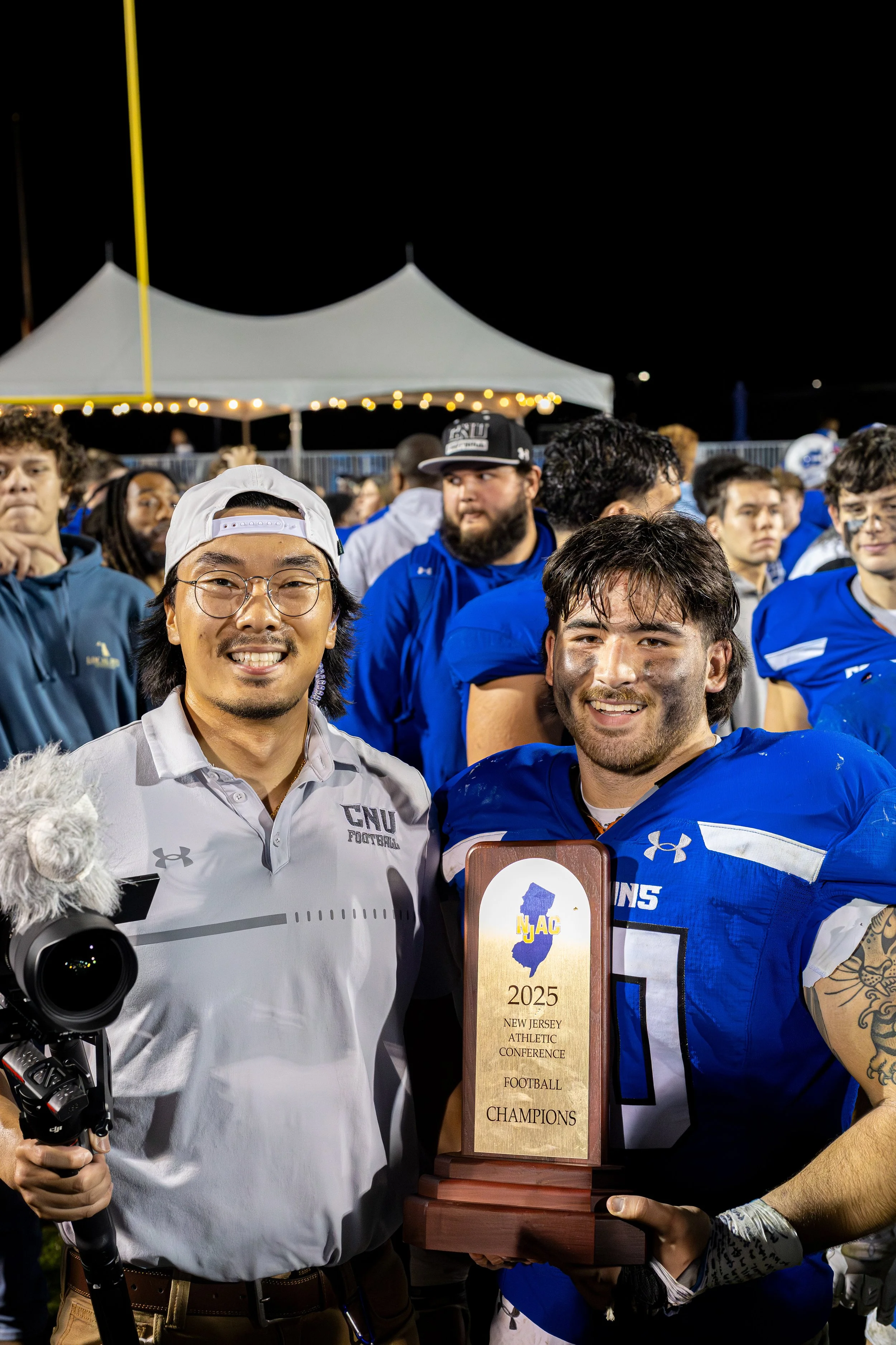 Two football players and a coach smiling while holding a championship trophy at night, with a crowd and a white tent in the background.