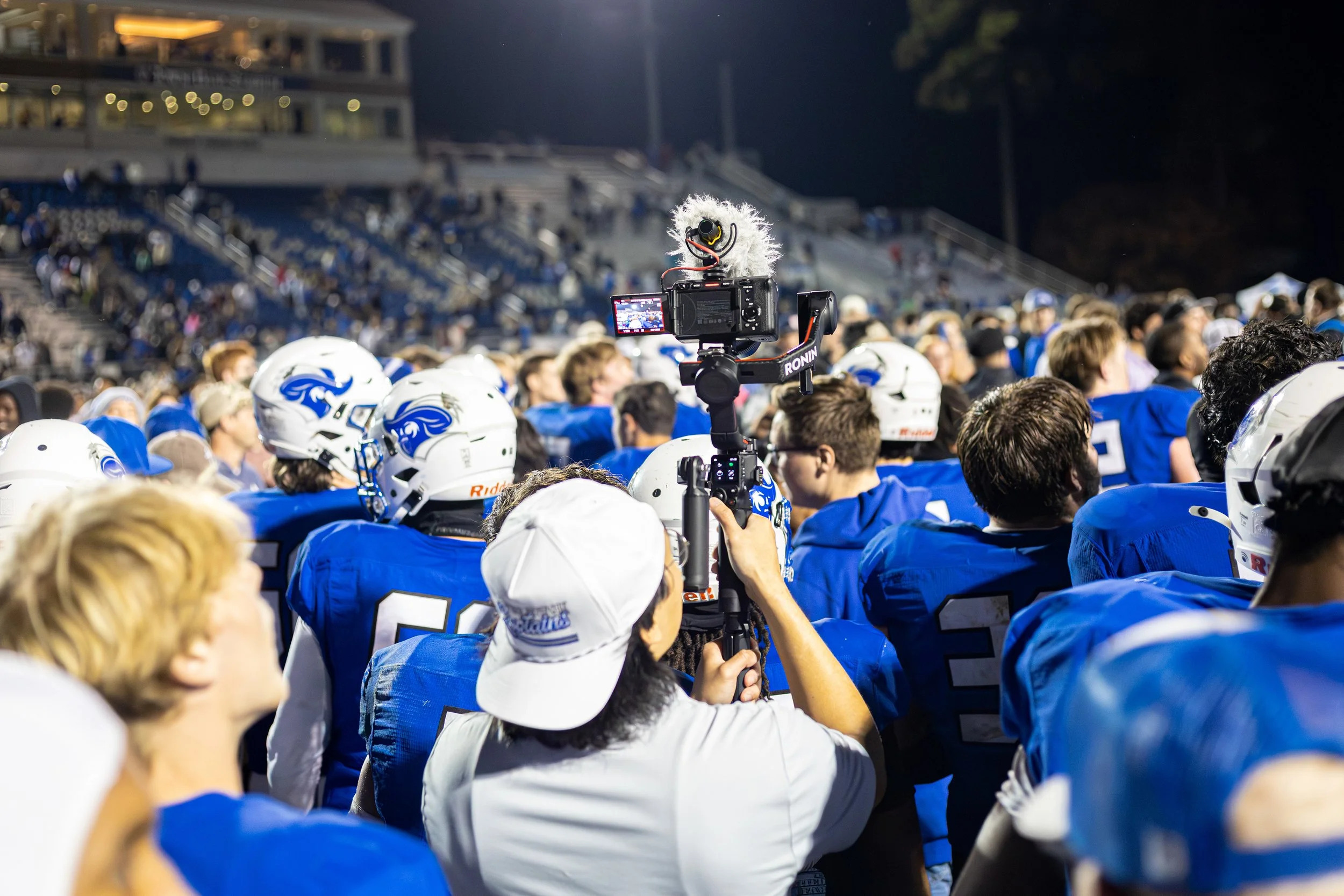 Football players in blue uniforms and white helmets gather on the field during a game or event at night, with a person filming the scene using a camera mounted on a stabilizer.