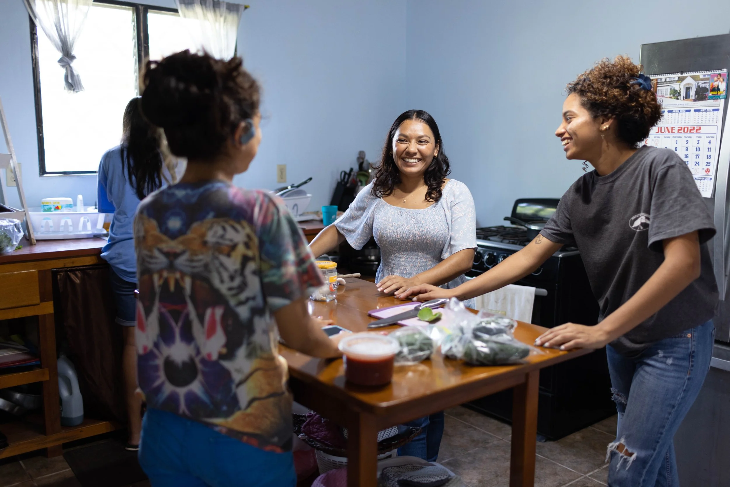 Four women socializing in a kitchen, preparing food and smiling. One woman is wearing a grey shirt, another in a colorful patterned shirt, and two others are partially visible. There are ingredients and containers on the wooden table.