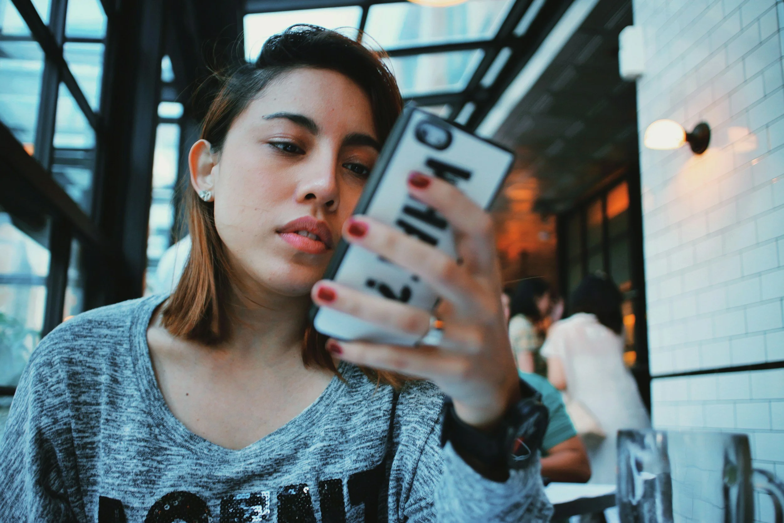 Young woman with brown hair and silver earrings looking at her phone inside a modern building with glass panels and white brick wall.