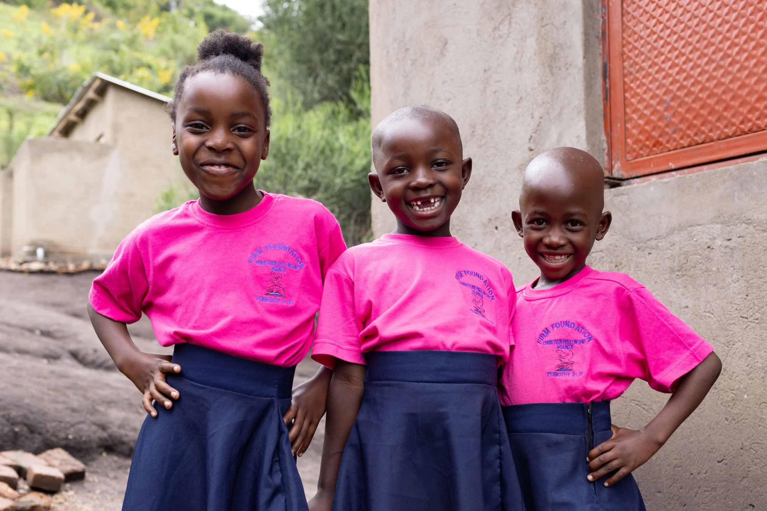 Three young girls with dark skin and short hair, wearing pink shirts and dark skirts, standing outdoors in front of a building with a red window and greenery in the background.