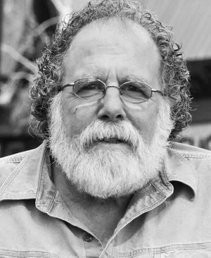 A black and white portrait of an older man with a beard and curly hair, wearing glasses and a collared shirt.