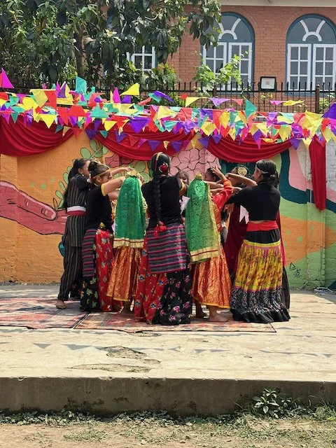 Group of people performing a traditional dance on an outdoor stage decorated with colorful bunting and a painted backdrop.